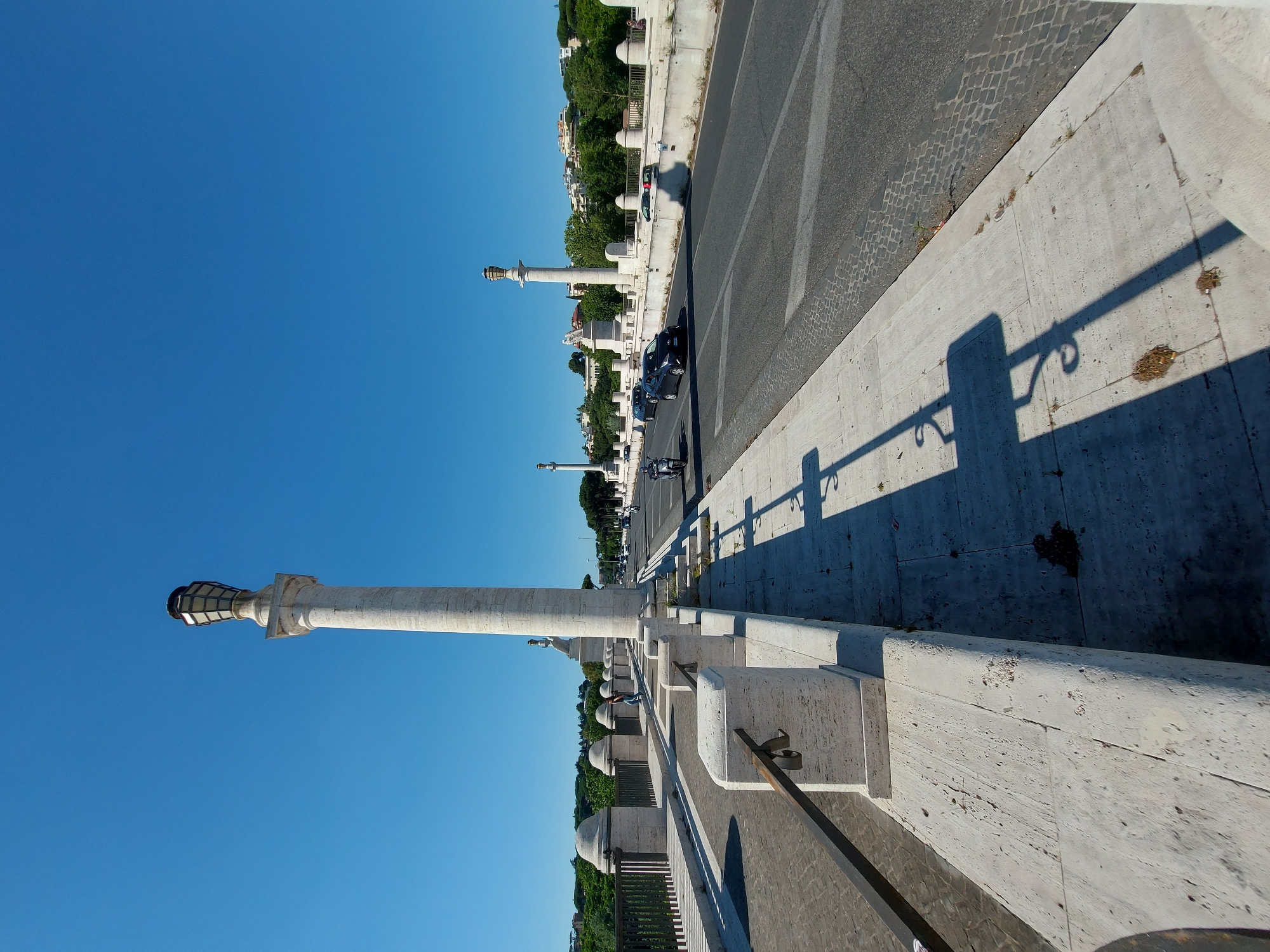 The image shows a sunny street scene in Rome, Italy. The camera is positioned on an elevated walkway, capturing a view of the street below. Along the walkway are decorative stone posts topped with light fixtures. The light fixtures extend horizontally into the clear blue sky. Below, the street is paved with cobblestones and asphalt. Cars and motorcycles are visible. The other side of the street features similar stone structures and vegetation.