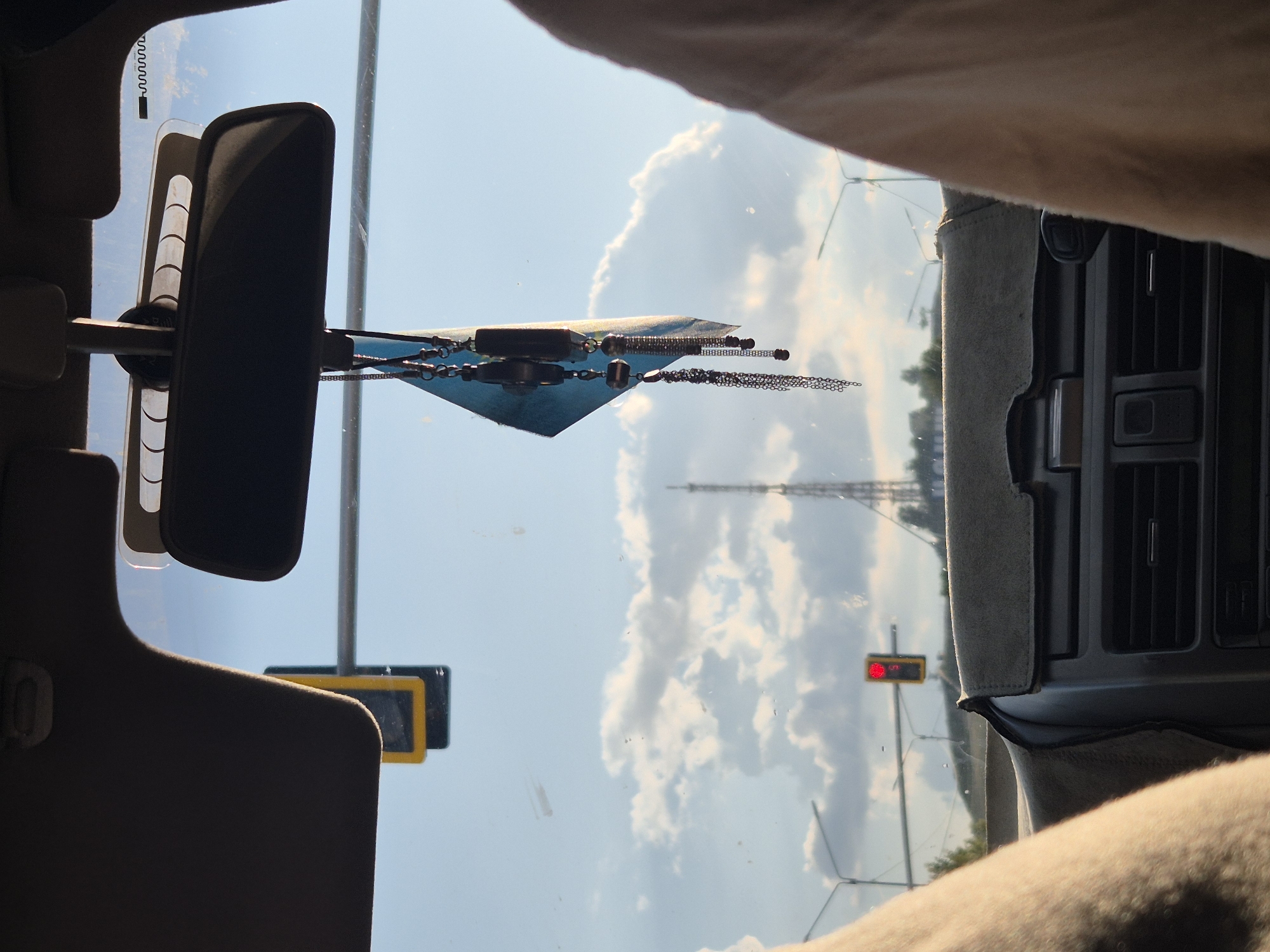 The image is a view from inside a car in Kokshetau, Kazakhstan, looking through the windshield. A rearview mirror is visible, with a hanging air freshener featuring a blue flag and metallic charms. Sunlight illuminates scattered clouds in a blue sky. In the distance, a radio tower stands against the horizon, with traffic lights indicating a red signal. The interior of the car shows part of the dashboard and the tops of seats.