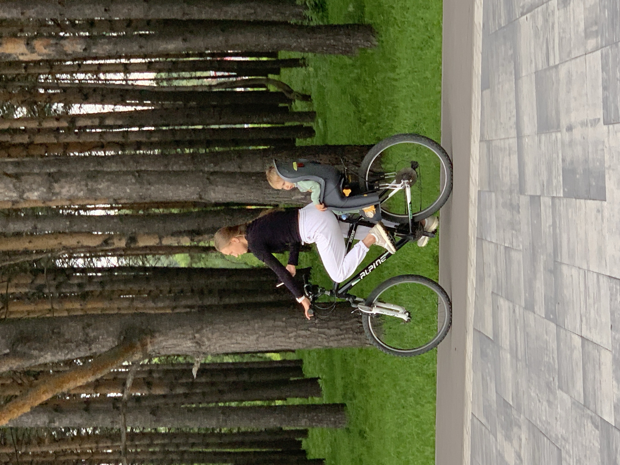 A young woman rides a bicycle with a child in a car seat on the back, along a paved path in a park. The woman is wearing a black long-sleeved shirt and white pants, and the child is wearing a light green outfit. They are surrounded by tall, slender trees with a grassy area in the background. The paved path is made of grey rectangular stones. The scene appears to be in a park within the city of Prokopyevsk, Russia.