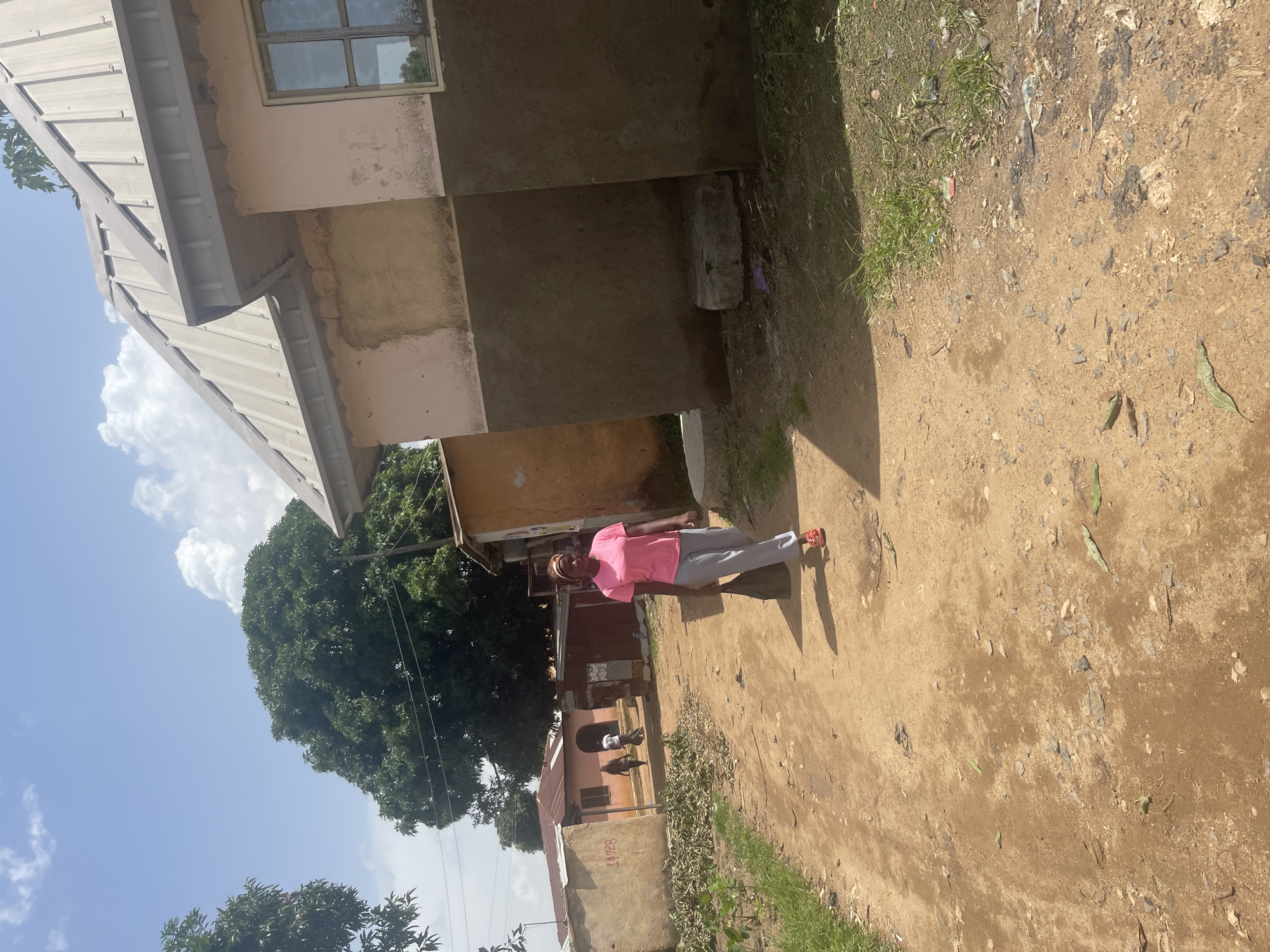 In Rafin Par, Nigeria, a woman walks down a dirt road, carrying a black bag in her right hand. She wears a pink t-shirt and grey pants, with red sandals on her feet. The road is lined with buildings constructed from concrete blocks and corrugated metal, some appearing weathered. Vegetation, including trees and grass, grows along the sides of the road, with some debris scattered on the ground. The sky is blue with scattered white clouds. The scene is illuminated by bright sunlight, casting shadows on the ground.