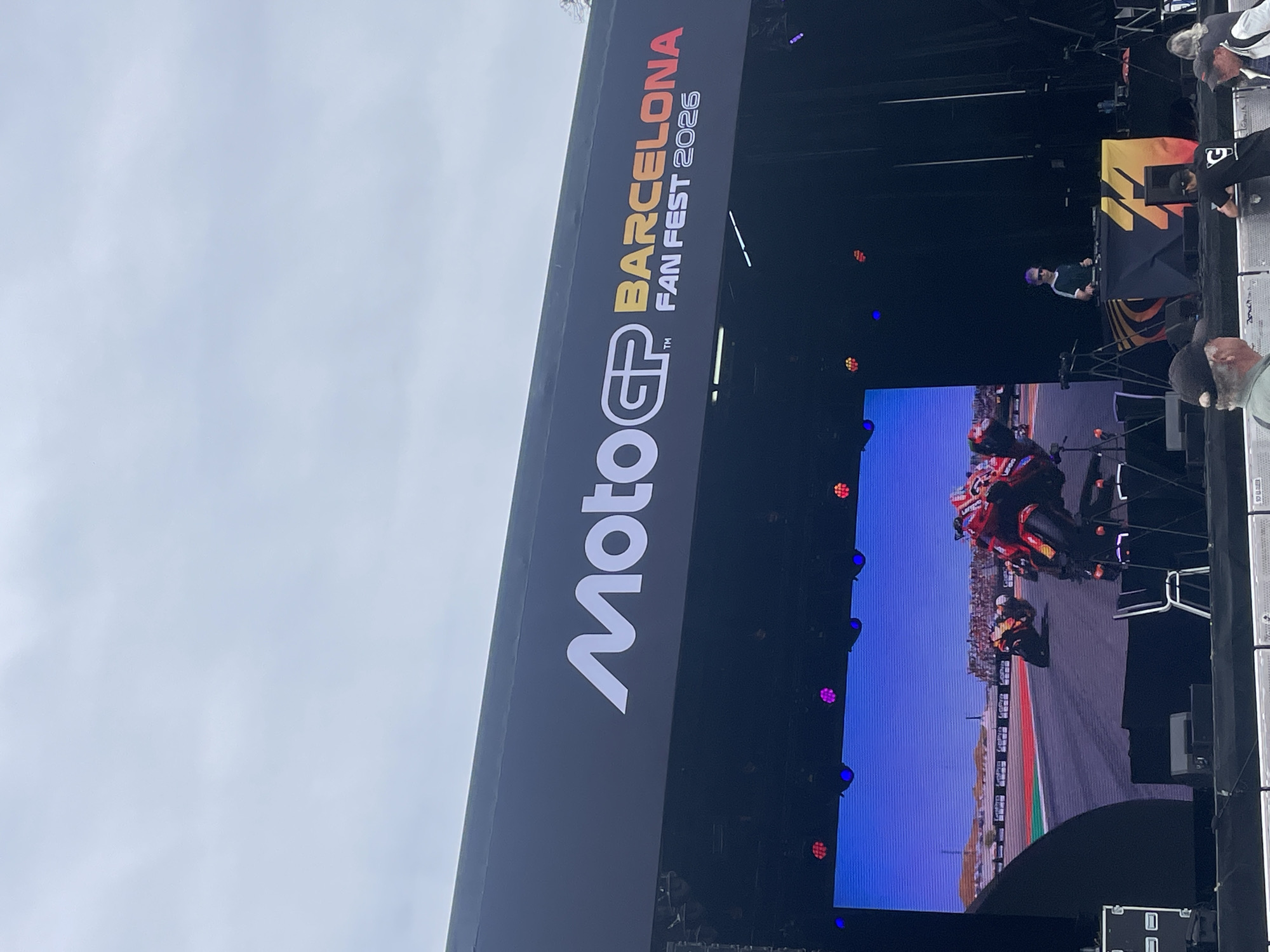 The image shows a stage set up for the MotoGP Barcelona Fan Fest 2026. A large screen displays a motorcycle race, while a DJ is visible on a higher platform. A crowd of people, some wearing hats, are positioned to the right of the stage. The sky is overcast.