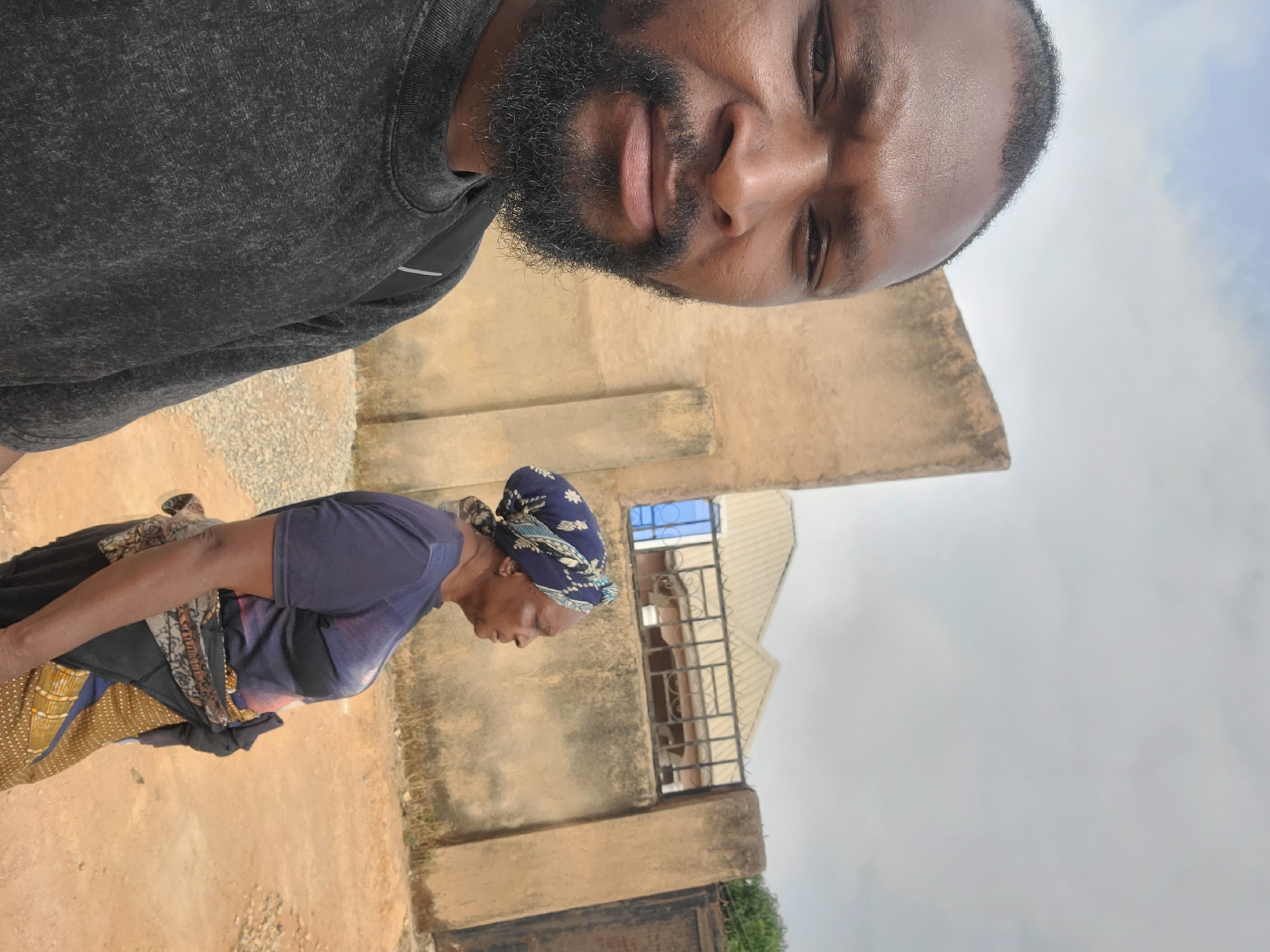 The image depicts a man in a dark grey top, presumably taking a selfie, with an older woman walking past in the background. The woman is wearing a blue top, a patterned skirt, and a headscarf. They are standing outside a beige building, with a window visible in the background. The sky is overcast. It is unclear from the image what state the scene is located in.