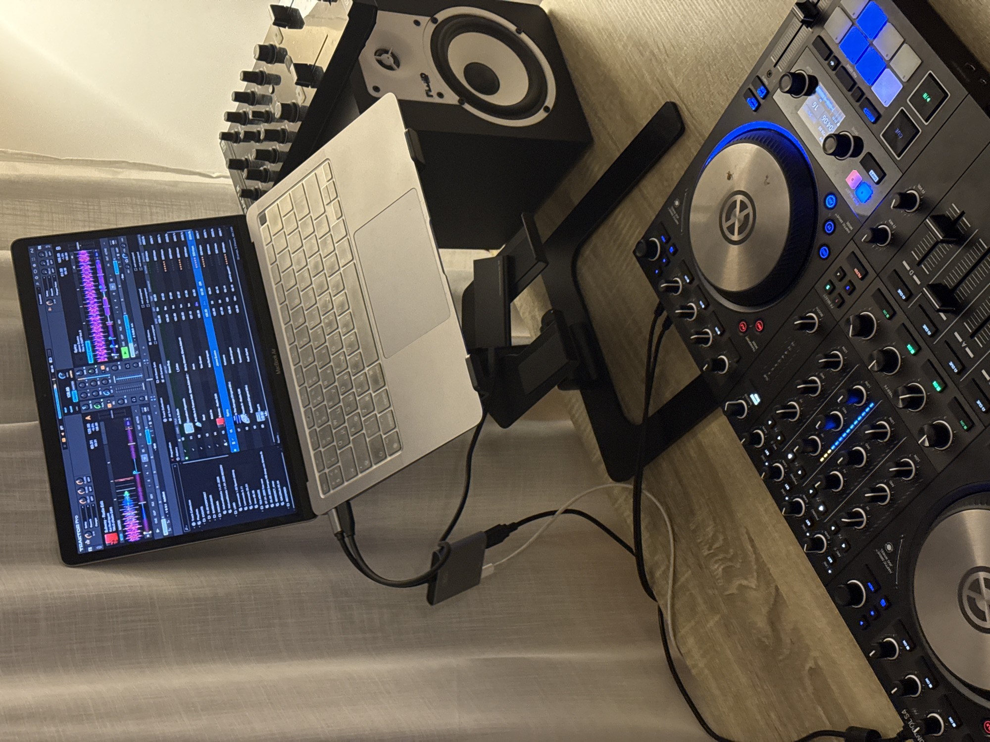 The image shows a DJ setup consisting of a laptop, DJ controller, speaker, and other equipment. The laptop screen displays the Traktor Pro DJ software, and the DJ controller has various knobs, buttons, and platters. The speaker is black with a white cone. The laptop is a MacBook Air. The environment appears to be a room with a curtain and a wooden surface. There is no indication that this is located in Boreti, Montenegro.