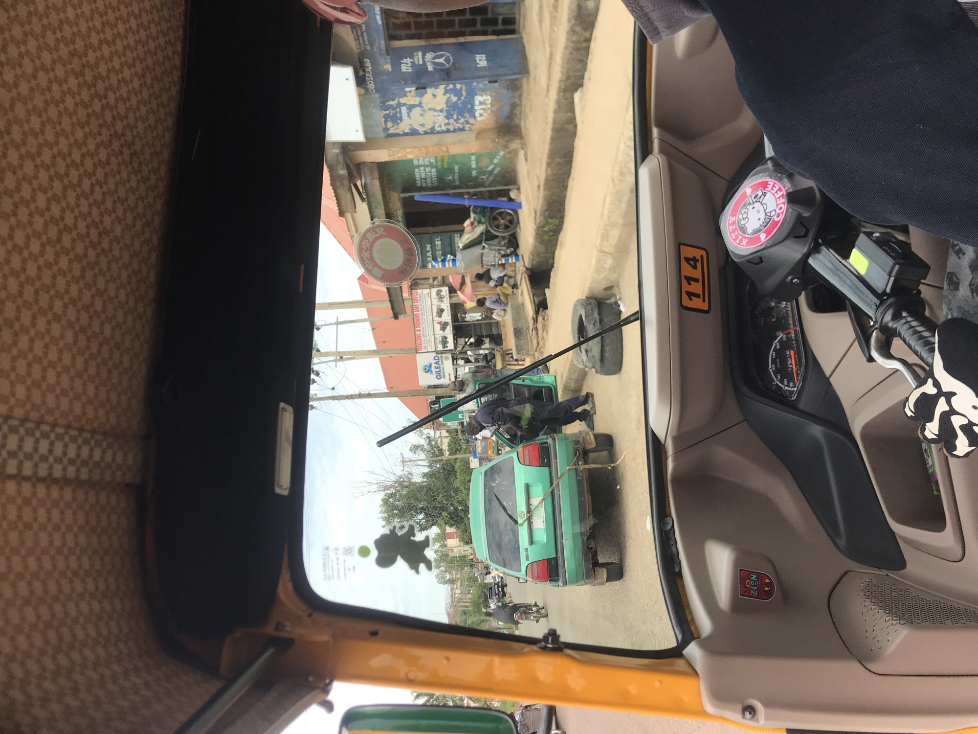 The image depicts a street scene in Rafin Par, Nigeria, viewed from the interior of a yellow and brown three-wheeled vehicle (tricycle). The tricycle's ceiling features a brown checkerboard pattern. The dashboard area includes a white rectangular placard with "114" in black, and a speedometer/gauge cluster. A circular red sticker with "KITTY COFFEE" and a cartoon cat is affixed to the handlebars. A "ZION" sticker is visible on the interior side panel. The driver's left arm, wearing a dark blue sleeve and a patterned white and black glove, is partially visible in the lower right foreground.
Through the front windshield, an elephant-shaped air freshener hangs, along with a red and white circular sticker displaying Asian characters. The street in front is an asphalt road. A light green, older model Volkswagen Golf hatchback is stationary in the middle of the lane, facing away from the viewer. Its rear bumper appears damaged, and a thin, light-colored rope or stick is tied to it, hanging down. The rear wiper is broken and angled upwards. Two men are standing near the rear of the green Volkswagen; one, wearing a dark uniform, is positioned at the car's open passenger-side door, while another man in a lighter shirt stands nearby. A long, dark pole is leaning against the car or lying on the ground next to it.
Further down the street, a motorcycle with a rider is visible in the left lane, moving away. On the right side of the road, a series of single-story buildings with corrugated metal roofs line the street. One building with a red roof displays a sign for "GILEAD MEDICAL CENTER". Adjacent to it, another building with a green facade has a sign partially visible, reading "BRANCH OFFICE OF NEPA JOS OPPOSITE STATION RD." Next to this is a dilapidated blue building with "1924" and "1213" painted on its exterior, alongside a Mercedes-Benz logo. Several individuals are visible near these buildings, some sitting on the ground or by a cart. An open concrete drainage ditch runs parallel to the road. Discarded tires and a tall, blue cylindrical object are present on the roadside. Utility poles with overhead wires are visible throughout the scene. The sky is overcast.