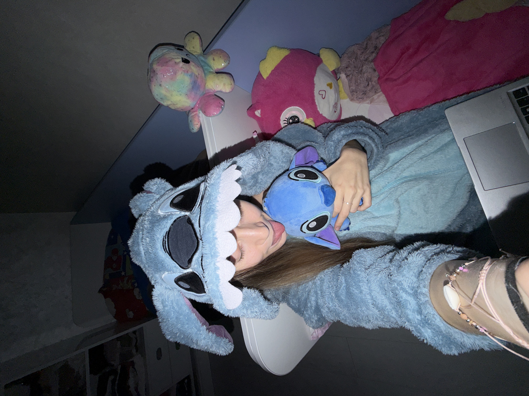 A person wearing a blue, fluffy "Stitch" onesie and a "Stitch" character hood is lying on their back, holding a smaller "Stitch" plush toy. They are sticking their tongue out. Above them are a rainbow octopus plush toy and a pink fish plush toy. A laptop is partially visible to their right, along with a pink blanket. The scene appears to be indoors, possibly in a bedroom. The location is unknown, and there is no information to suggest it is in the city of Plovdiv, Bulgaria.