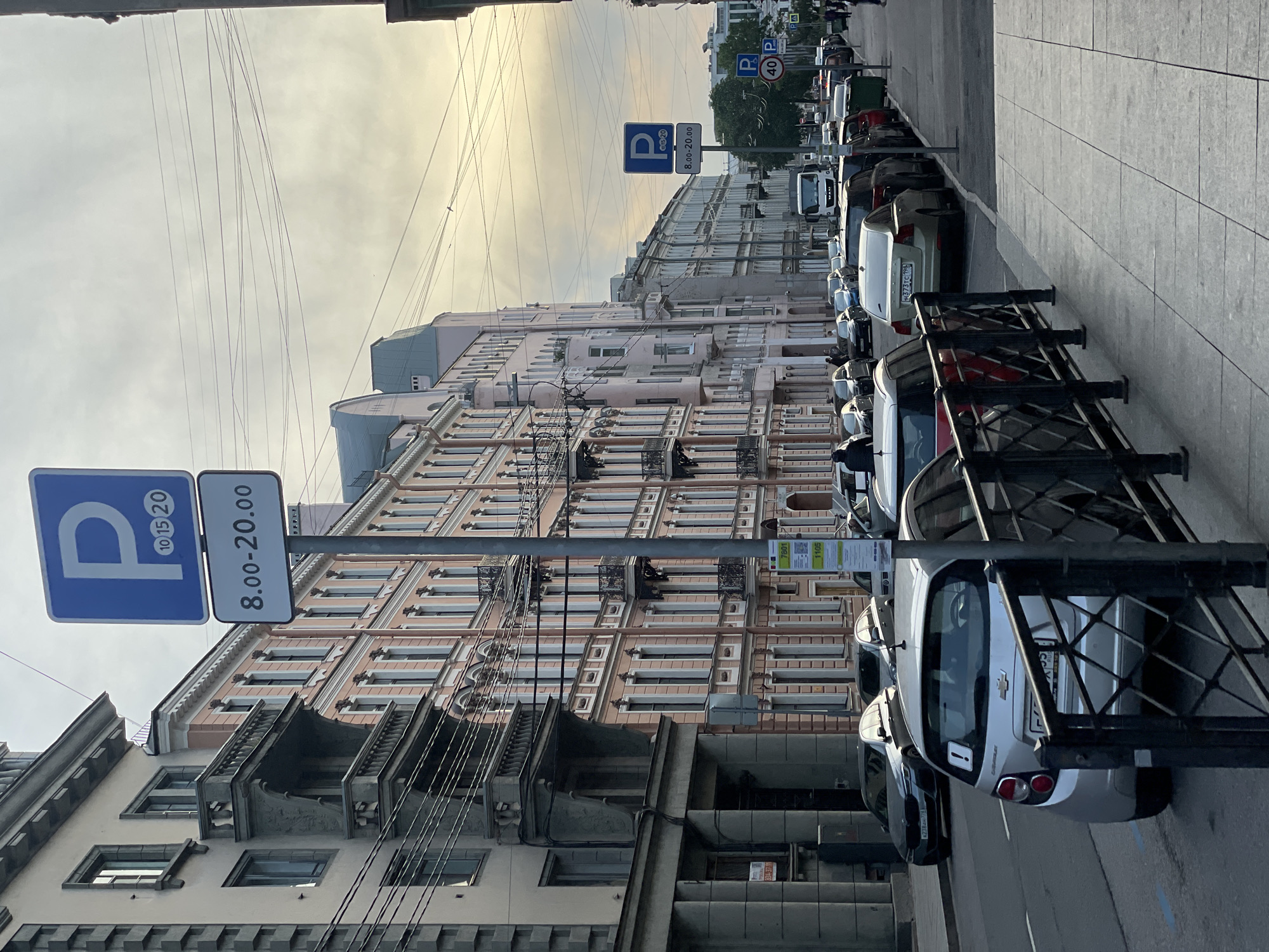 This image captures a street scene in the city of Saint Petersburg, Russia. The perspective is from a sidewalk, looking down a street lined with multi-story historic buildings.
On the left, a five-story building features a light beige or pale grey facade with darker, possibly stone, architectural details and prominent, ornate balconies spanning multiple floors. Utility wires crisscross in front of this building.
Dominating the central and right portions of the image is a long, multi-story building characterized by a distinct salmon-pink or peach-colored facade with white decorative window frames and numerous small, wrought-iron balconies. Further down the street, additional buildings with similar architectural styles, but lighter in color, recede into the distance. Visible rooftops include pitched designs, some with dormers, and one with a teal-colored roofline. Overhead utility lines are prominent against the overcast, slightly hazy sky.
In the foreground, a black metal barrier separates the sidewalk from the street. Numerous cars are parked bumper-to-bumper along both sides of the street, indicated by blue markings on the asphalt near the curb. A silver Chevrolet Aveo hatchback, bearing a white exclamation mark sticker on its rear window and a Russian license plate (E477OP 193 RUS), is prominently parked near the foreground barrier. Further down, a light-colored sedan with a Russian license plate (H373TC 198 RUS, indicating the Saint Petersburg region) is visible, alongside other parked vehicles including a silver BMW (A238AM 777 RUS) and a red car. A white MAN truck is visible further down the street on the right side.
Two blue rectangular 'P' (parking) signs are visible on tall poles. The sign in the foreground clearly displays "10 15 20" within the 'P' symbol, and below it, "8.00-20.00", indicating paid parking hours. A similar but less distinct parking sign is seen further down the street on the right, also with "8.00-20.00" and an additional sign below that appears to show "40" (possibly a speed limit or parking zone detail). A green and white information stand, likely for public transport or parking payment, displaying numbers "7801" and "1105", stands on the sidewalk near the center-right.
Several pedestrians are sparsely distributed on the sidewalks, walking or standing, but no specific actions or interactions are discernible. The sidewalk is paved with square concrete slabs. The overall lighting suggests either early morning or late afternoon with soft, diffused sunlight.