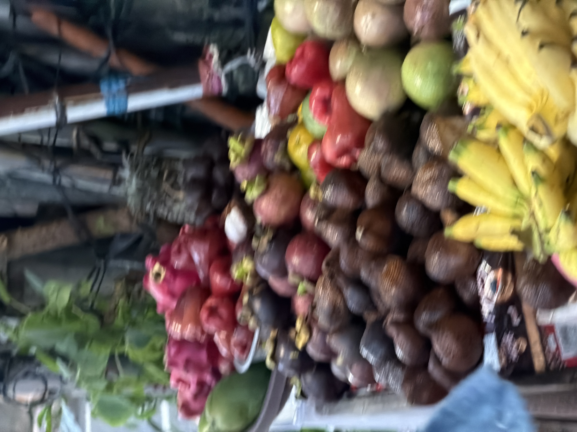 An outdoor fruit stall in the city of Ubud District, Indonesia, displays a variety of fresh produce. In the foreground, bright yellow bananas are visible, some with green tips. Behind them, a jumble of fruits includes dark, scaled fruits, likely salak, and various round fruits in shades of red, green, and light brown, possibly rose apples, rambutan, or mangosteen. A large green fruit, possibly a mango, is situated to the left, behind a pile of vibrant pink and red fruits, which could be dragon fruit or lychee. The background is blurred but suggests a natural or rustic setting with greenery and wooden structures.