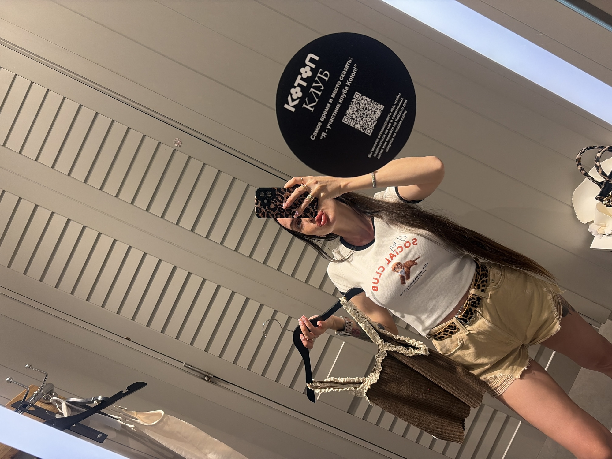 A young woman is taking a mirror selfie in a fitting room. She is wearing a white t-shirt with "Social Club" printed on it in red letters and a graphic of a dog. She is also wearing light brown denim shorts with a leopard print belt. She is holding a brown knitted top with lace trim on a hanger. The fitting room has beige slatted walls and a white door. A circular black sticker with Russian text and a QR code is visible on the wall. The text on the sticker appears to be promotional material for "Koton Club." The overall impression is that this is a fitting room in a Koton clothing store. Based on the text on the sticker, the store is likely located in Saint Petersburg, Russia.