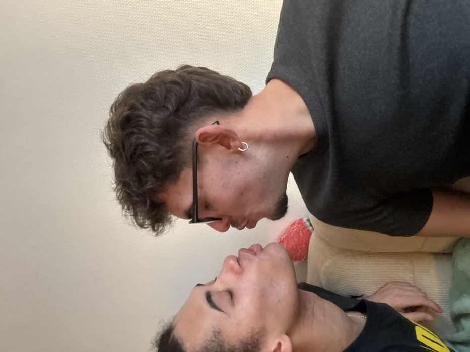 Two people, likely men, are in close proximity. One man stands, slightly angled, wearing glasses, a gray shirt, and a silver hoop earring, looking downward. The other man lies down, looking upward. Their faces are near touching, as if about to kiss. They are indoors with a visible couch and wall.