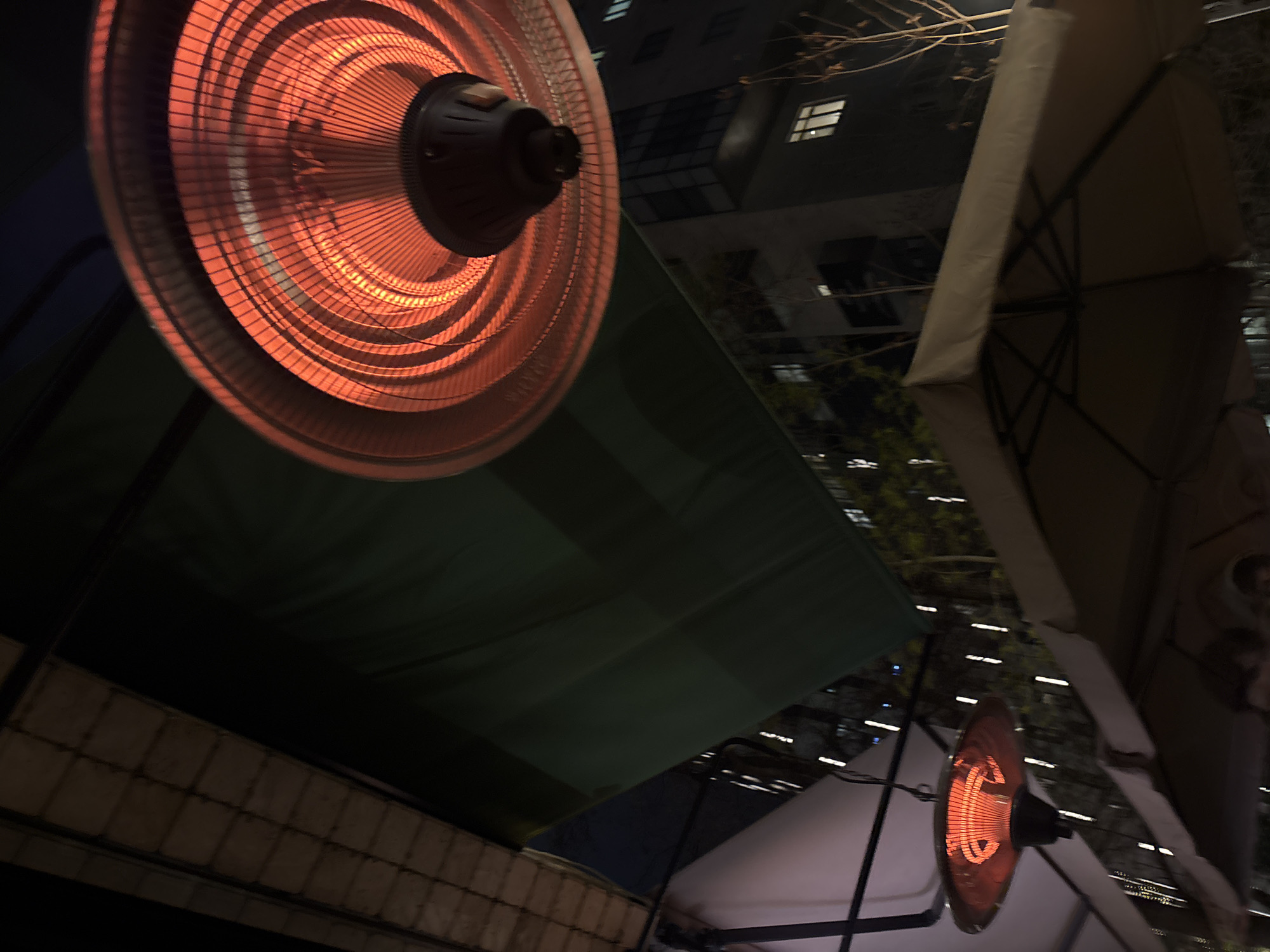 This nighttime image, set in an urban environment in Yerevan, Armenia, captures an outdoor area illuminated by two glowing electric patio heaters. The prominent heater in the upper left is a circular, infrared type, emitting a bright orange-red light from its heating element, protected by a metallic grill. A smaller, similarly glowing heater is visible in the lower right.
These heaters are suspended, likely providing warmth for an outdoor seating area, parts of which are covered by awnings or large umbrellas. A dark green awning stretches across the middle of the frame, while sections of lighter-colored umbrellas with their support structures are visible on the right. The presence of the heaters strongly suggests cool or cold weather conditions.
In the background, against a dark sky, a building with multiple illuminated windows indicates a city setting. Bare tree branches are also discernible against the dim light, adding to the evening atmosphere. The overall scene is dark, with the heaters and distant building lights serving as primary sources of illumination. No people or visible text are present in the image. The perspective is angled upwards, emphasizing the overhead heaters and the urban backdrop.