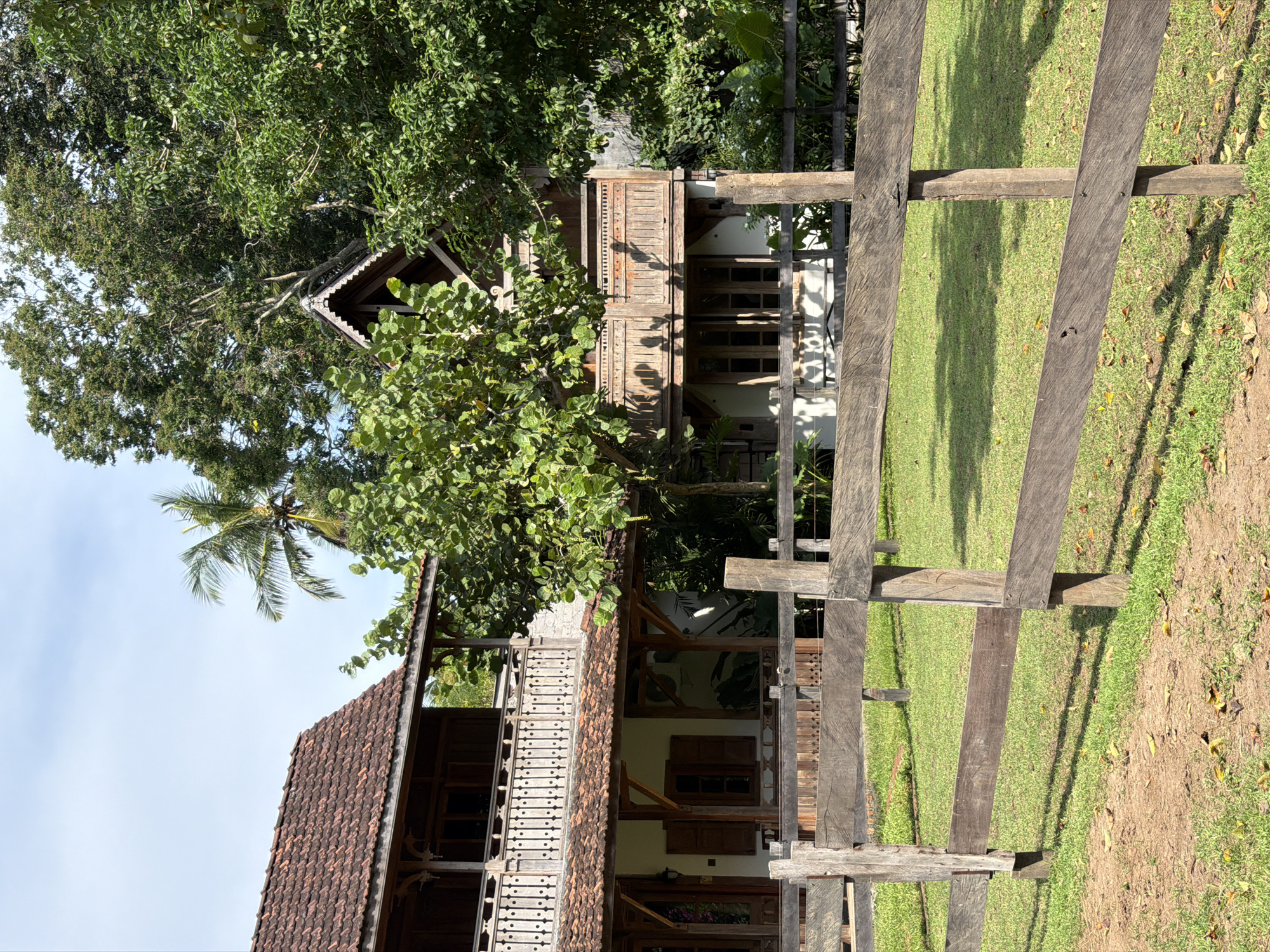 The image shows a traditional wooden building with a tiled roof and intricate wooden carvings, located in a grassy area in Kediri, Indonesia. A wooden fence runs along the foreground, casting shadows on the green lawn. The sky is partly visible through the trees.