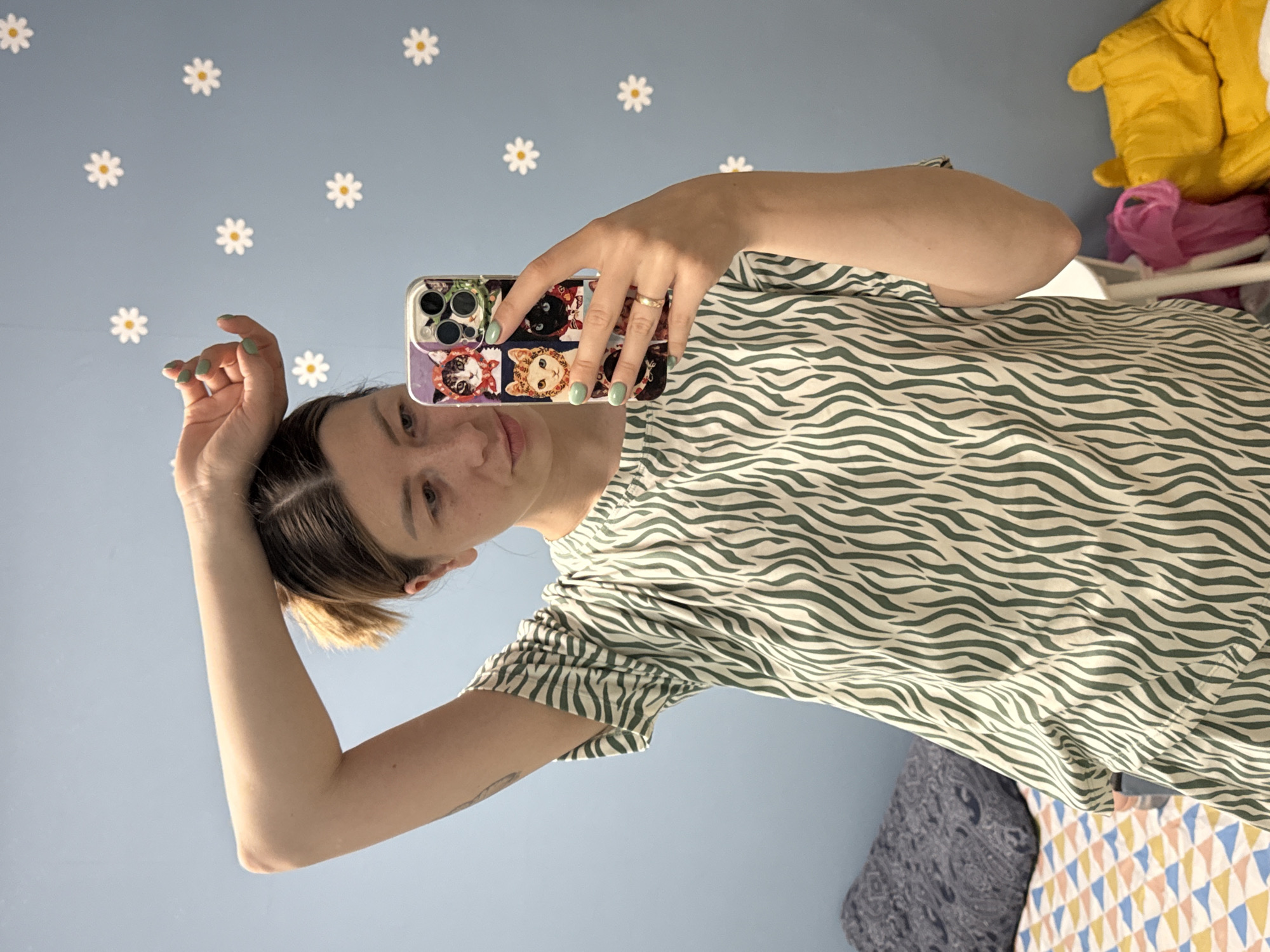 A woman is taking a selfie in what appears to be a room with a blue wall decorated with white daisy stickers. She is wearing a green and white zebra-striped shirt and holding a phone with a cat-themed case. Her nails are painted light green, and she has a ring on her finger. The background shows parts of a bed with patterned linens and some clothing items, including a yellow jacket. The location is not identifiable as Moscow, Russia from the provided image.