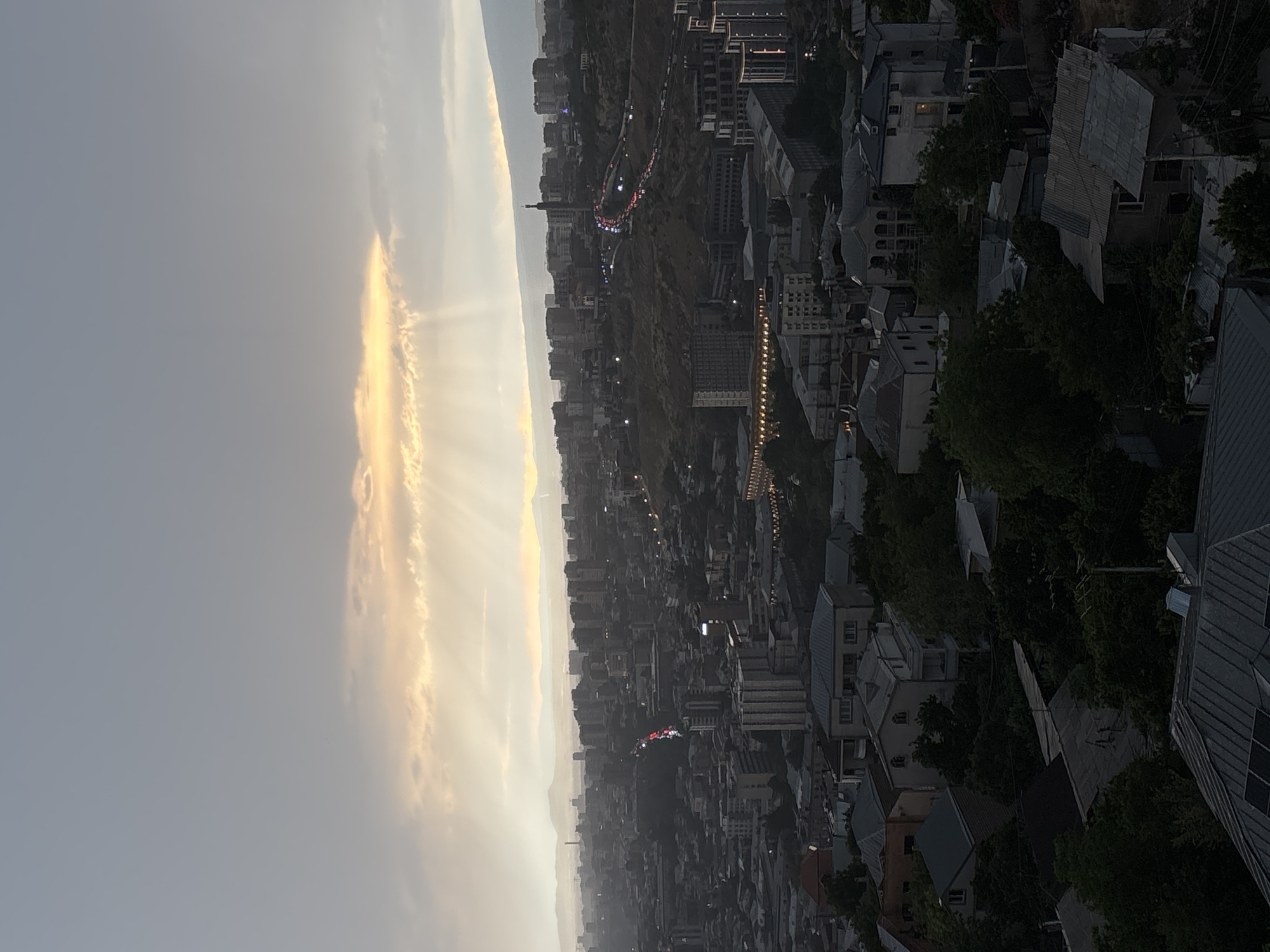 This image captures a wide, elevated view of Yerevan, Armenia, at dusk or dawn. The sky is dramatic, with a band of clouds illuminated by the sun, casting crepuscular rays downwards. A dense urban landscape stretches out below, characterized by a mix of apartment buildings and smaller residential houses. In the mid-ground, a busy highway, likely the Northern Avenue or a similar major thoroughfare, is visible with car headlights and taillights creating streaks of red and white light. The terrain of the city appears hilly, with buildings constructed on slopes. Distant mountains are faintly visible on the horizon, softened by the atmospheric conditions. The overall impression is of a sprawling city under a moody sky. There are no visible people or specific actions depicted in the scene.
