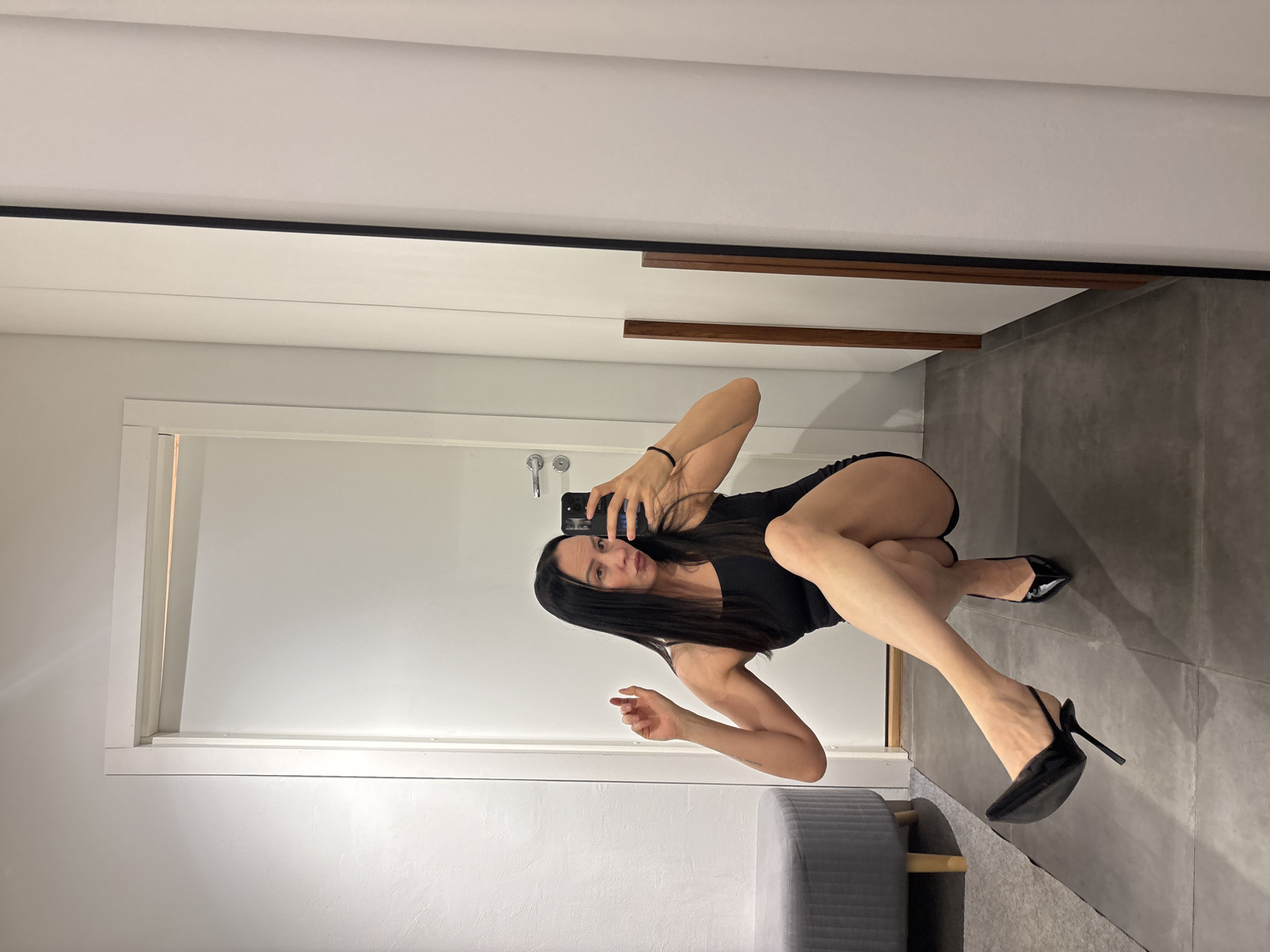 A woman is taking a mirror selfie indoors. She is wearing a black dress and black high heels. She is crouching in front of a mirror in what appears to be a fitting room. The room has white walls and gray tile flooring. There is a gray ottoman in the foreground. Based on the metadata, the location is Chaikovsky, Russia.