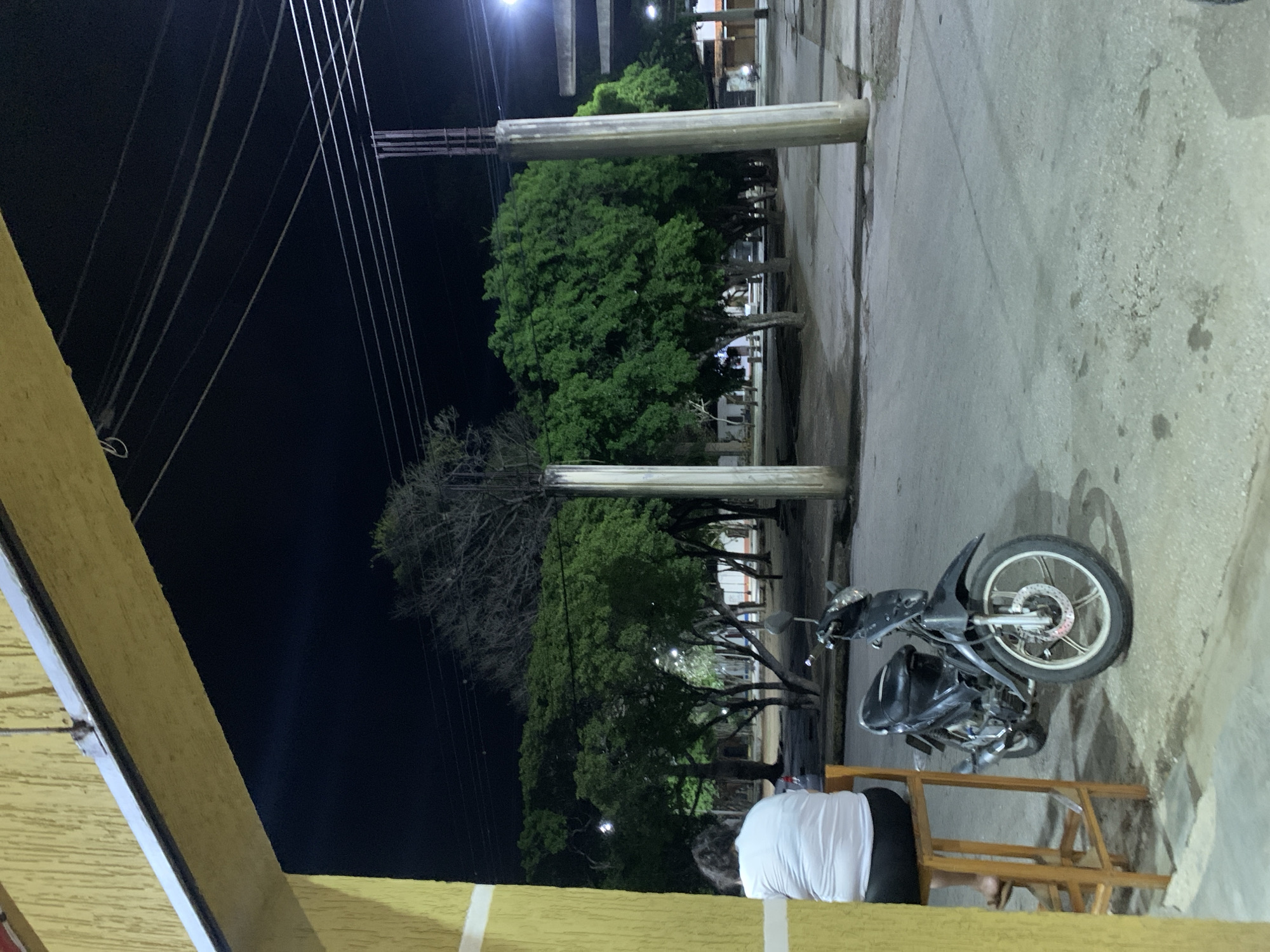 This nighttime image, taken in Santa Ana, Venezuela, captures a quiet street scene from an elevated perspective, likely a balcony or porch. The time of day is clearly night, with a dark sky illuminated only by distant streetlights and artificial light sources creating various glows and reflections.
In the immediate foreground, a yellow-painted wall or railing runs across the bottom and left edges of the frame. A person is visible from behind, sitting on a simple wooden stool or chair, wearing a white t-shirt and dark trousers. They appear to be looking out at the street.
The middle ground features a paved area, likely a sidewalk or a small street, on which a black motorcycle is parked on the right side. The motorcycle faces left, with its front wheel and disc brake prominently visible. Further into the scene, several large trees with dense green foliage line what appears to be a public space or park. Interspersed among these are some trees with sparser, bare branches. Two prominent concrete or utility poles stand vertically in the mid-ground, separating the paved area from the treeline.
The dark night sky in the upper left is crisscrossed by multiple overhead power lines or utility cables, extending from unseen poles on the left toward the right. The overall atmosphere of the scene is calm and tranquil, with no discernible dynamic activity or strong emotions, simply a quiet observation of an urban environment at night. No visible text is present in the image.