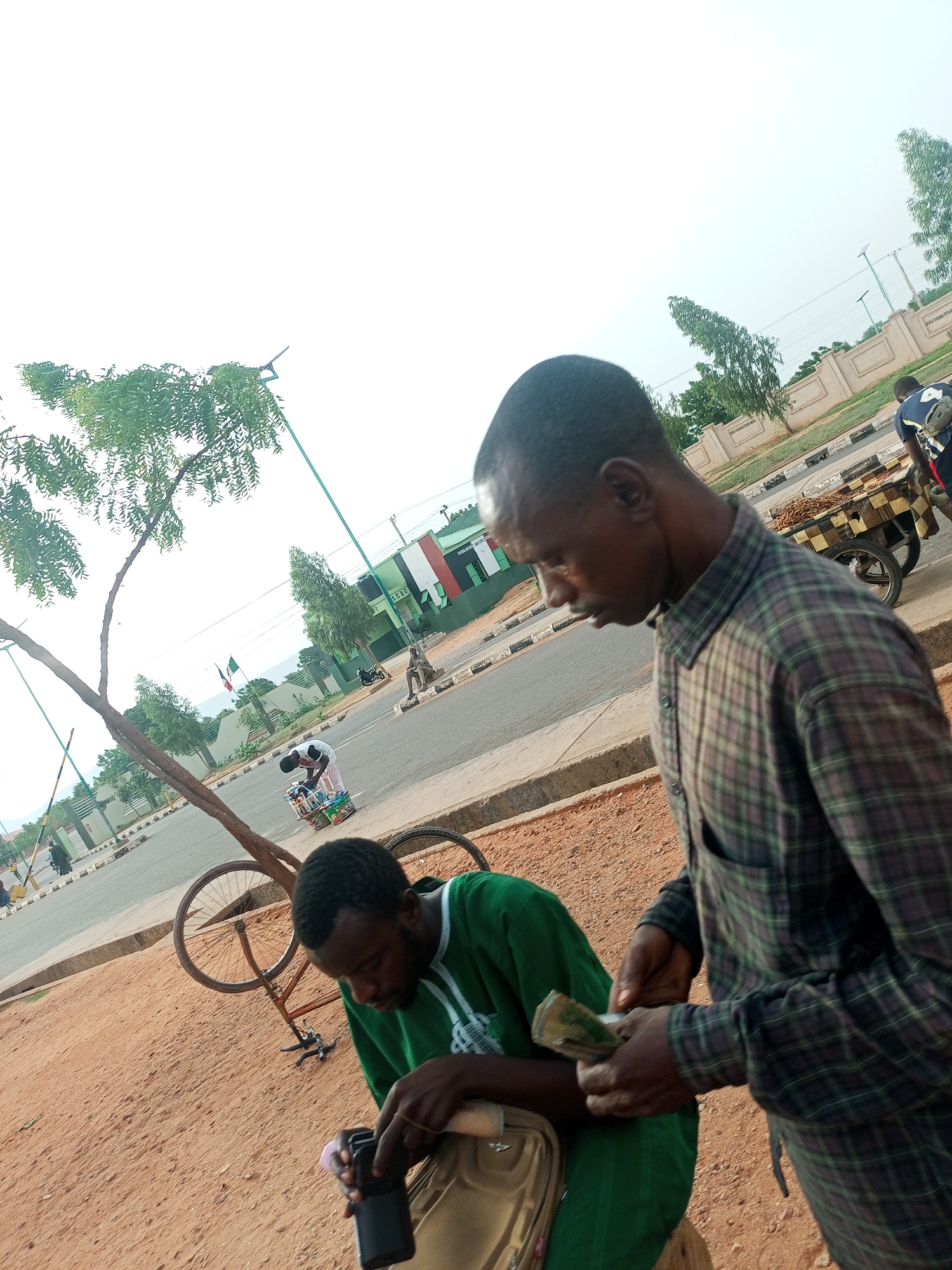 Two men are standing near a road in Gombe, Nigeria. One man, wearing a green shirt, is seated and appears to be holding a camera or a phone. The other man, wearing a plaid shirt, is holding and counting money. A broken bicycle is nearby, along with a tree on the side of the road. A cart laden with goods is being wheeled in the distance. In the background are buildings and a road with pedestrian and vehicular traffic.