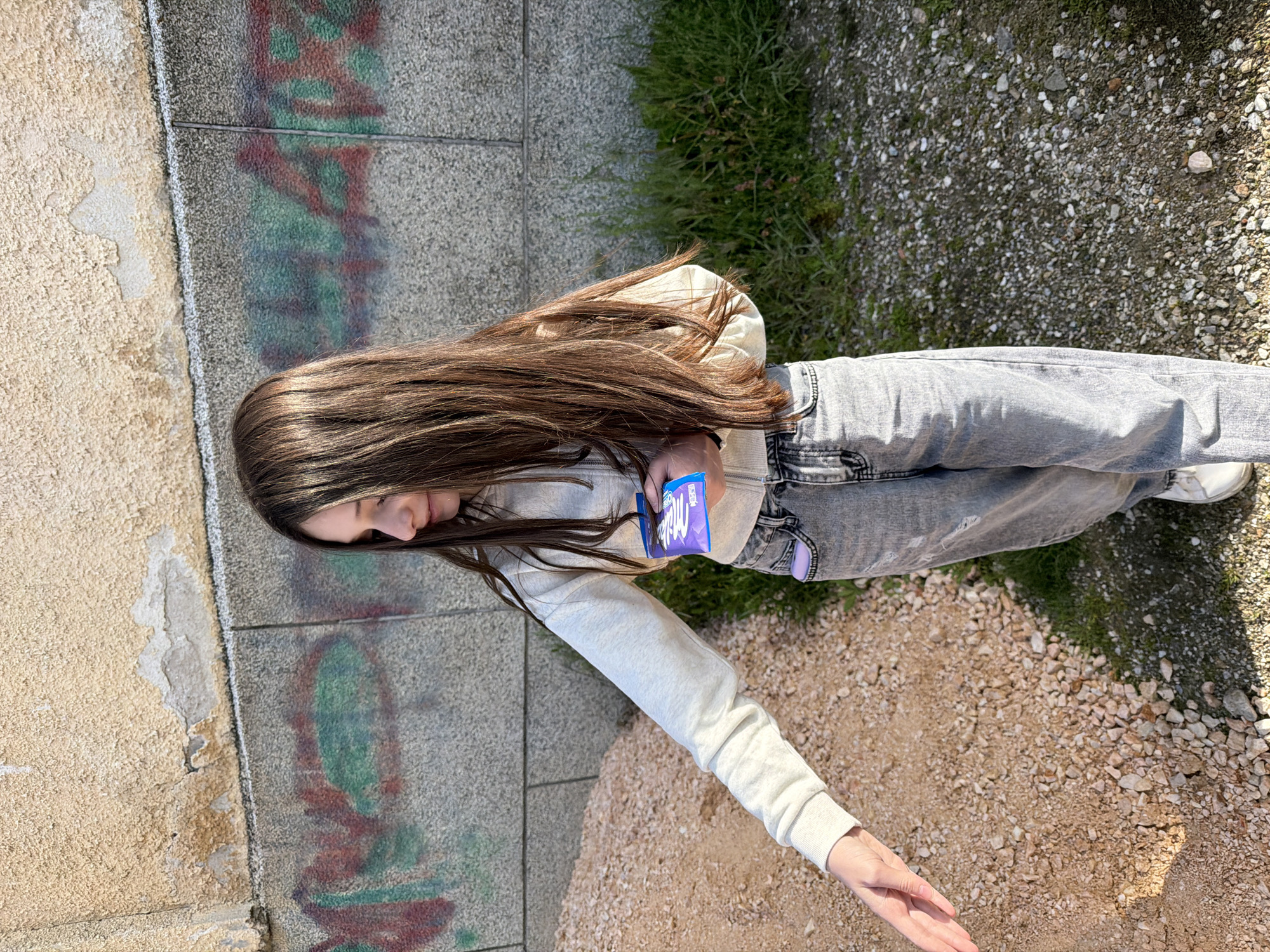 A girl is positioned diagonally, with her head towards the left edge and her feet pointing to the right edge. She has long, brown hair and is wearing a light gray hooded sweatshirt and faded gray jeans. She is holding a purple "Milka" chocolate bar. Her left arm is extended towards the bottom right corner. The background includes a concrete wall on the left with some graffiti and discoloration, and ground covered with small rocks and some patches of green grass or weeds on the right. The image is taken outdoors in Zmeitsa, Bulgaria.