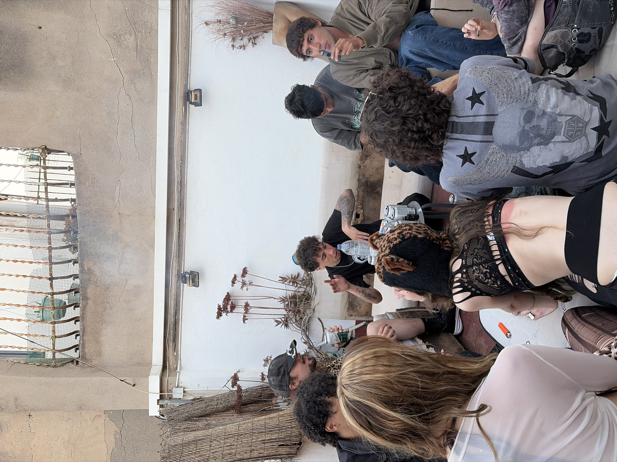 A group of approximately nine individuals are gathered on a rooftop in Barcelona, Spain. They are seated and appear to be engaged in conversation. Some of them are dressed in casual clothing, and one individual is wearing a top with a skull design. There is an assortment of objects scattered around, including water bottles, a lighter, and what appear to be discarded cans. The rooftop is partially enclosed by a white wall and a metal railing, with a view of the city visible in the background. Dry plants and woven bamboo elements are used for decoration. The overall scene suggests a casual gathering.