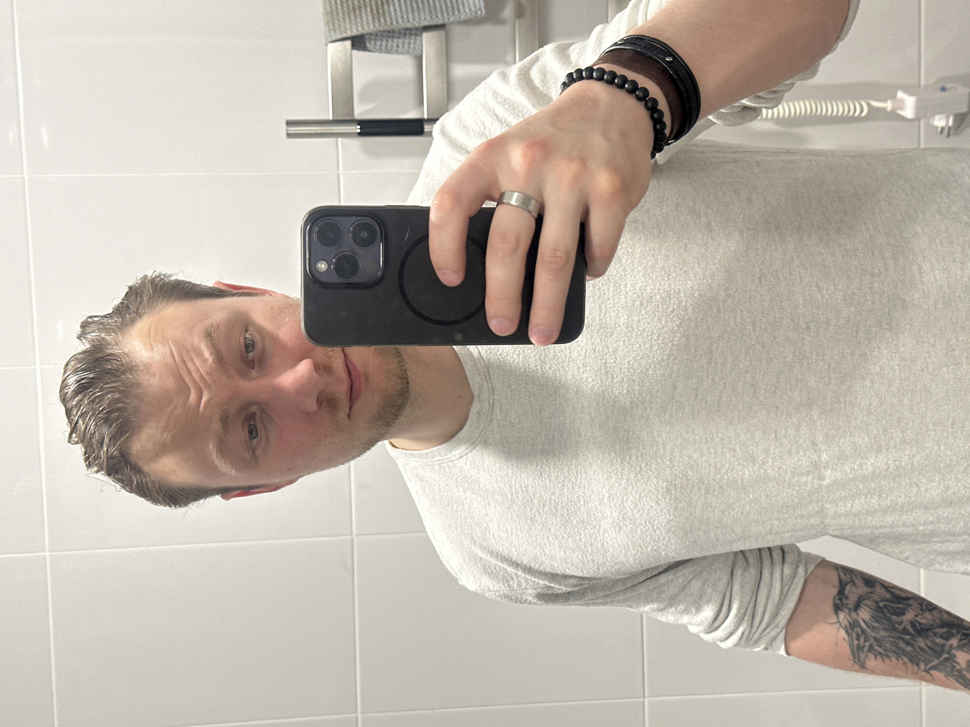 A man is taking a selfie in a bathroom. He is holding a black smartphone in his right hand, which is covering part of his face. He is wearing a white t-shirt and has a tattoo on his left forearm. On his right wrist, he has several bracelets. He is wearing a ring on his right hand. The bathroom has white tiled walls and a towel rack is visible in the background. The scene is not explicitly linked to Sochi, Russia.