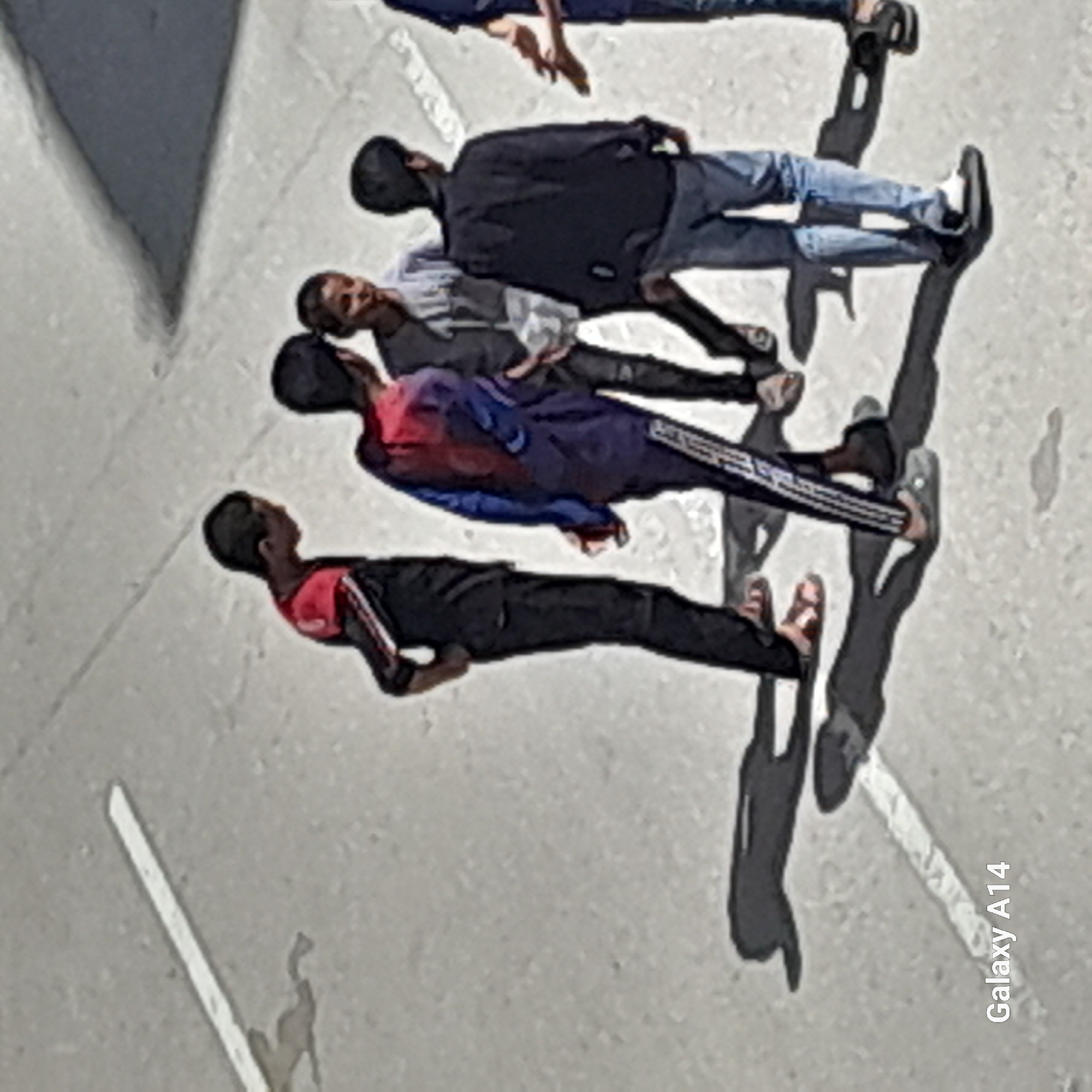 The image appears to be taken from an elevated perspective, showing a group of four individuals lying on a concrete surface, likely a street or sidewalk. They are positioned in a line, each person casting a distinct shadow that extends away from them. The concrete surface has painted white lines indicating potential lanes or markings.
The individuals are dressed in varied casual attire. One person is wearing a dark-colored shirt and blue jeans, while another is wearing a light-colored shirt. The remaining two are wearing black, blue, and red color block outfits. Their positioning and attire suggest they might be relaxing or performing a coordinated activity.
The overall environment appears to be an urban or suburban setting, with the concrete surface indicating a street or open space. The details about the specific city and location are unknown.