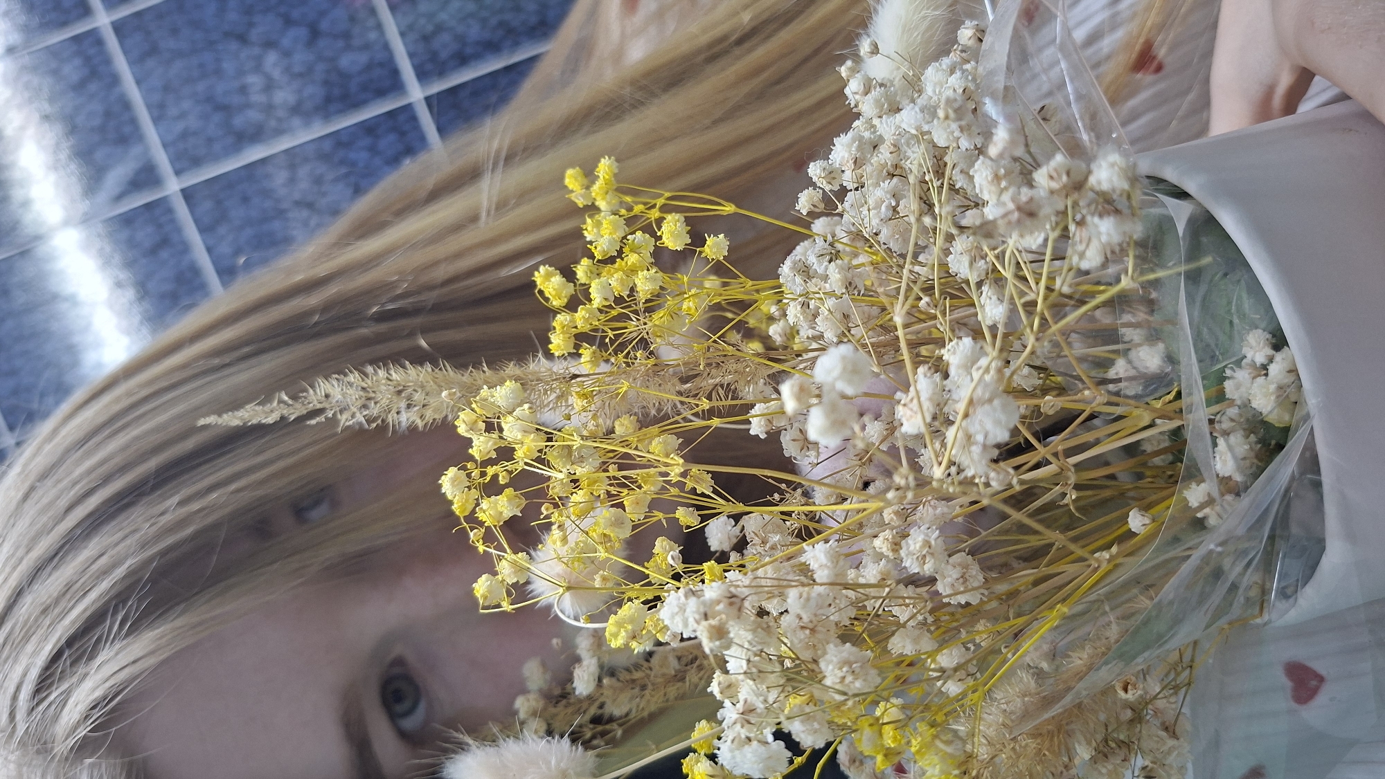 A close-up, rotated, slightly blurred image captures a partial view of a person with light blonde hair, partially obscuring the face, specifically the area around the left eye. The hair appears to be layered and falls to the side. A bouquet of dried flowers, consisting of small white and yellow blooms, dominates the foreground, partially covering the person’s face. The bouquet is wrapped in clear plastic. In the background, a grid-like pattern suggests tiled flooring or a wall. The location is Chaikovsky, Russia.