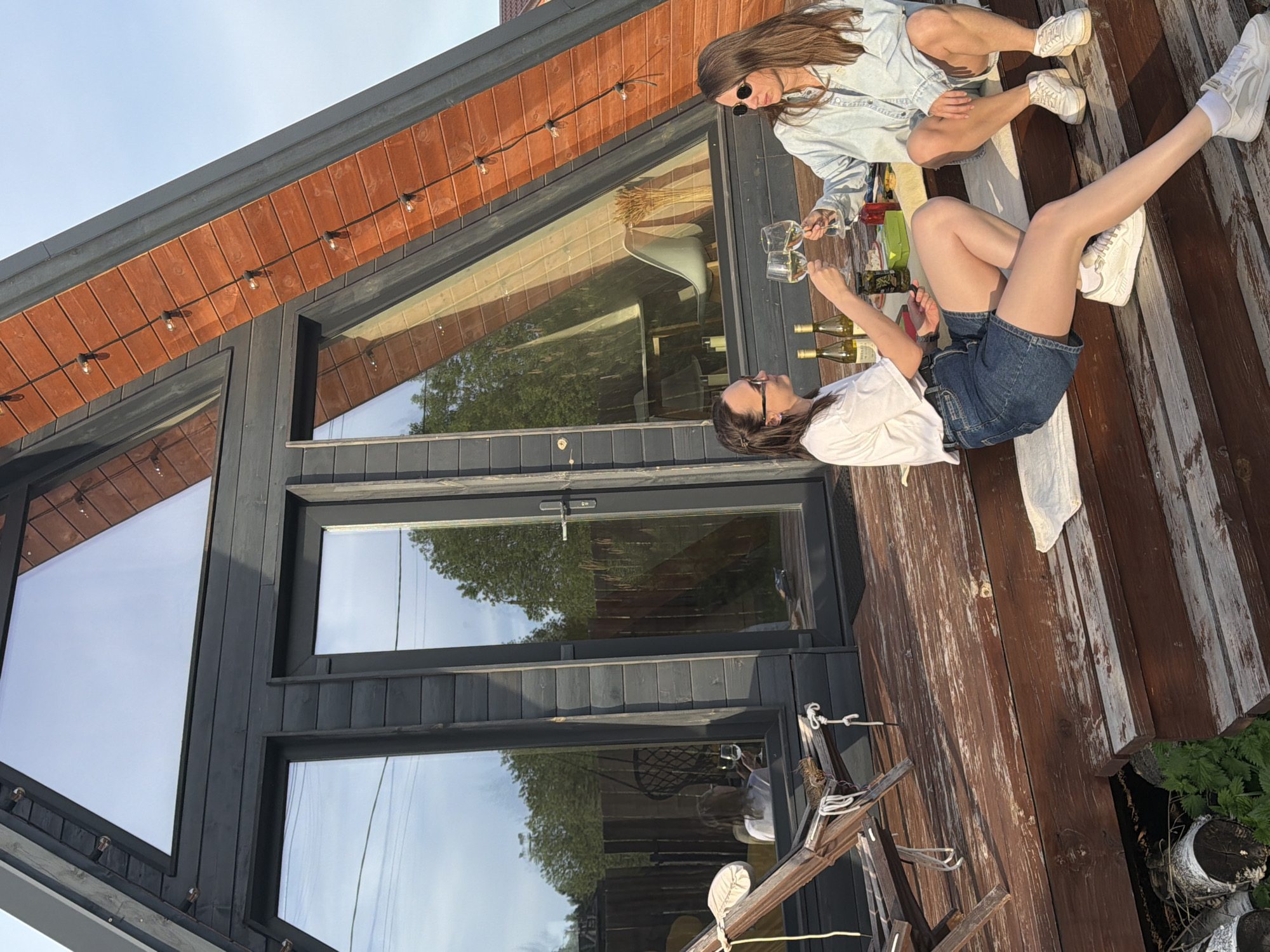 Two women are sitting on a wooden deck next to a modern cabin, possibly in Prikamskii, Russia, based on the prompt. The woman on the left is wearing a white shirt, denim shorts, and sneakers, and she is holding a wine glass. The woman on the right is wearing a denim jacket, shorts, and sneakers, and she is also holding a wine glass. There are several bottles of wine on the deck between them. The cabin has large windows that reflect the surrounding trees. There is a string of lights along the top of the cabin.