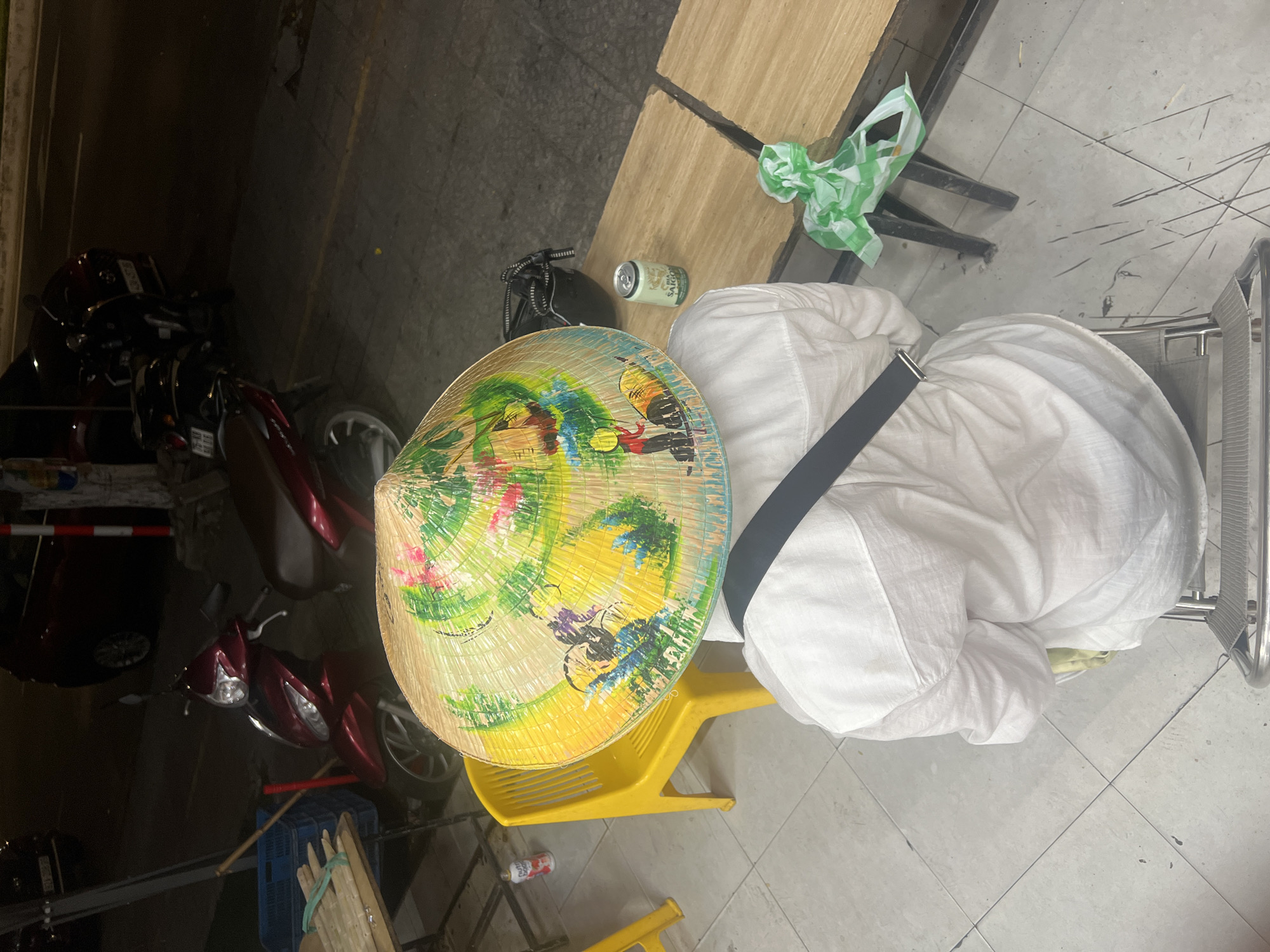 This is a photograph taken at night in a city environment. A person wearing a white shirt and a conical hat painted with various colors is seated on a metal chair. The hat features imagery that appears to depict rural scenes with figures and animals. The person is viewed from behind and slightly from above.
In the background, there are several motorcycles parked on a paved area. A red car is partially visible to the left. A bright yellow plastic chair is positioned to the left of the seated person. A table with some items on it, including what looks like bundles of sugarcane and a blue crate, is also visible on the left side of the image. To the right of the seated person, there is a wooden bench with a silver can of beer and a crumpled green and white plastic bag. The ground is tiled.
The scene is likely on a street or sidewalk, suggesting a casual setting such as a roadside eatery or a market stall. The presence of multiple motorcycles and the general urban atmosphere, coupled with the specific style of the conical hat and the beer brand (likely Vietnamese), strongly suggest the location is in Đà Nẵng, Vietnam, given the common association of these elements with street life in Vietnamese cities.