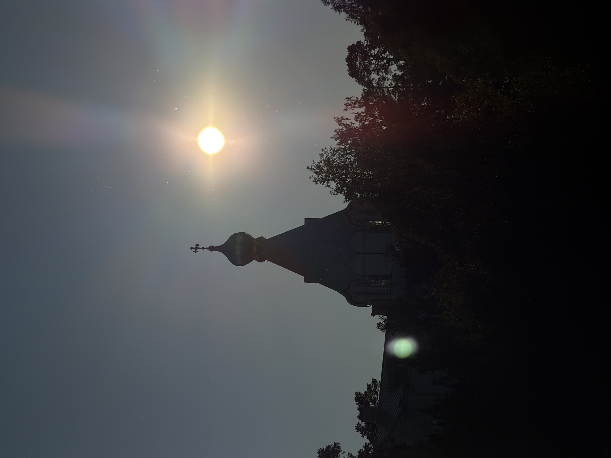 The image shows a silhouetted view of the Valaam Monastery, located on Valaam Island in the Republic of Karelia, Russia. The main subject is a prominent church dome topped with a cross, set against a twilight sky. The sun is a bright, almost blown-out orb in the upper right portion of the frame, casting a strong lens flare and visible light rays. Smaller celestial bodies, likely stars, are faintly visible against the darkening sky. The foreground is dominated by dark silhouettes of trees, with some foliage illuminated by the setting sun or moonlight, creating a dappled effect. To the left, a portion of another building, likely part of the monastery complex, is visible with a faint, greenish lens flare in front of it. The overall atmosphere is serene and somewhat mystical, characteristic of twilight or nighttime photography of religious sites. There are no people visible in the image.
