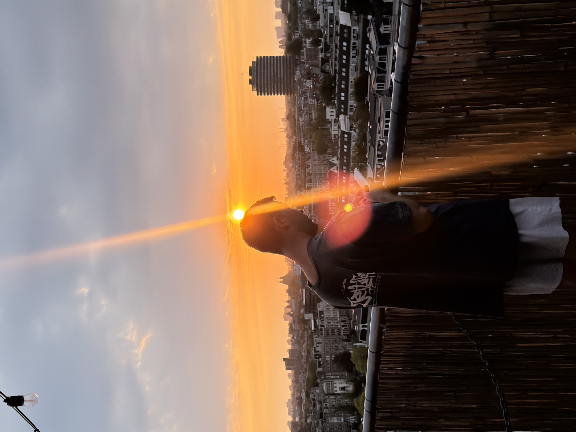 A person stands on a rooftop in Amsterdam, Netherlands, looking at their phone as the sun sets. The sky is filled with orange and yellow hues from the sunset, with some clouds visible. The person is wearing a dark t-shirt and light-colored shorts. In the background, the city skyline is visible, with various buildings and rooftops. A string of lights hangs from a pole in the upper left corner of the image. The rooftop has a bamboo fence. The sun's rays create a lens flare effect in the image.