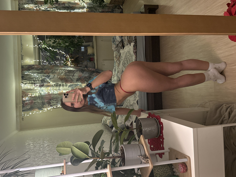 A woman in a tie-dye shirt, red thong, and white socks takes a mirror selfie. She holds a phone in her right hand. The background includes a bed, a window with lights, curtains, and a shelf with potted plants.