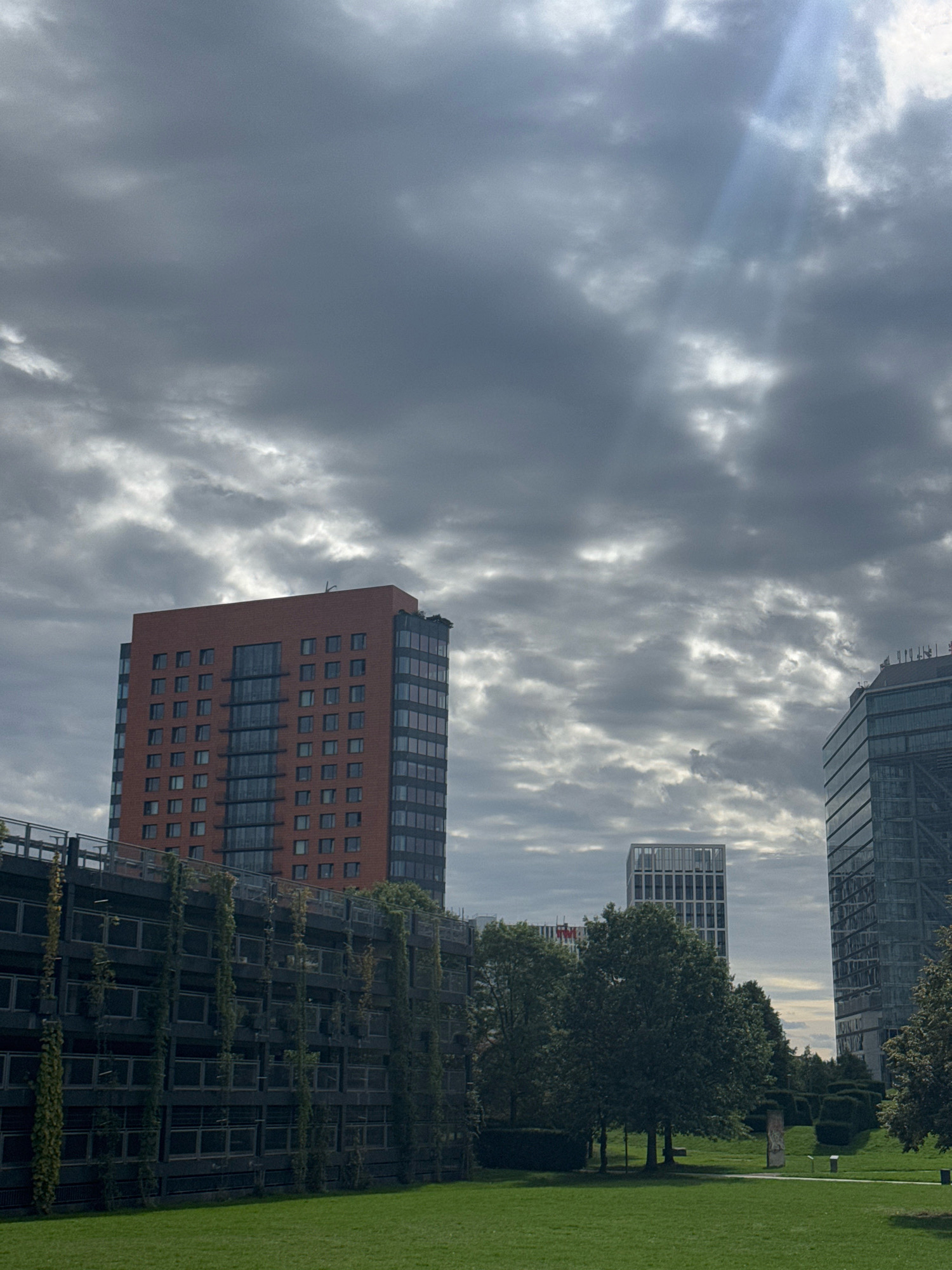 The image depicts a cloudy sky with shafts of sunlight breaking through, over a cityscape. Prominently featured is a tall, reddish-brown brick building with numerous windows, situated to the left of center. To its right, and slightly further back, is a modern glass skyscraper. In the foreground, a dark, multi-story structure covered in creeping vines is visible, with a lush green lawn in front of it. Several trees and manicured bushes are scattered across the grassy area. The scene is suggestive of a business district or urban park in Dusseldorf, Germany, possibly in an area like the Medienhafen. There are no people or discernible activity.
