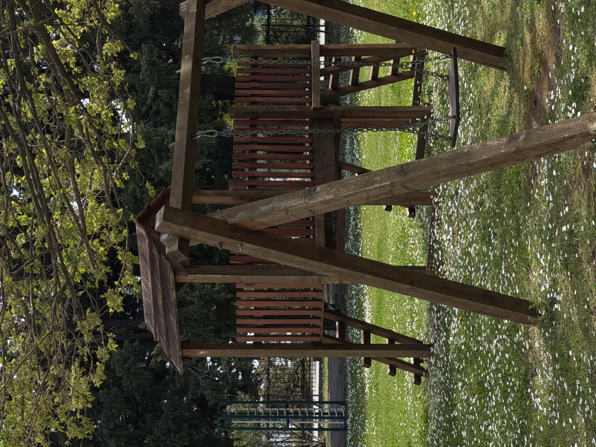 The image depicts a wooden playset located in an outdoor area, likely a park. The playset includes a two-level structure with ladder, swing and a set of stairs. The surrounding environment is a grassy area with small white flowers scattered throughout. The scene appears to be in the daylight. Some trees are visible in the background. It's difficult to pinpoint the exact location in Budva, Montenegro from the image alone.