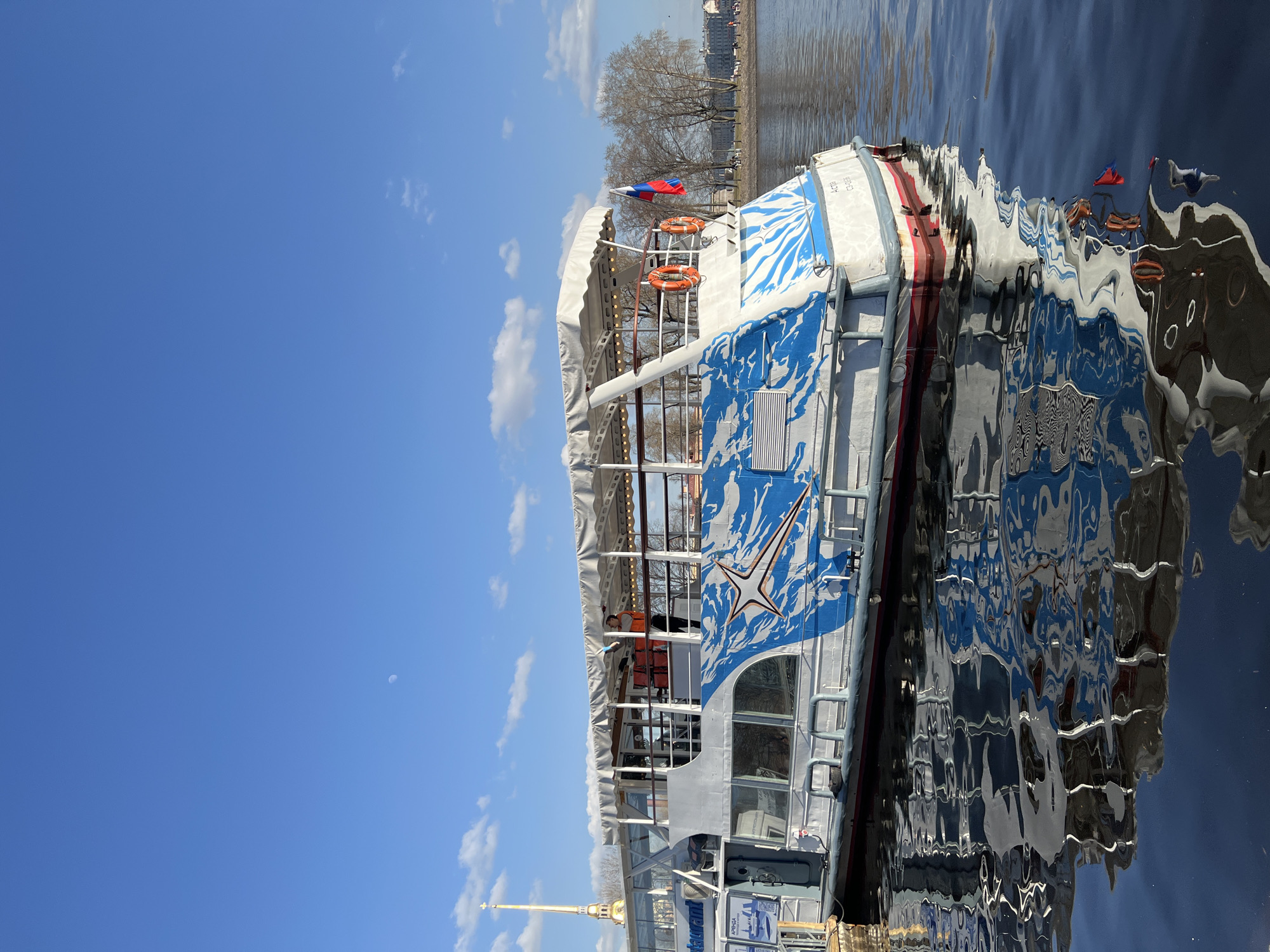The scene depicts a passenger boat docked on a body of water in Saint Petersburg, Russia, under a clear blue sky with sparse white clouds and a visible crescent moon. The golden spire of the Peter and Paul Cathedral is prominently visible in the background to the left.
The primary subject is a multi-deck white passenger boat, identified with the name "Астра" (Astra) and registration "СЗ-038" on its stern. Its hull features a large blue and white stylized graphic pattern resembling a star or splash. The boat has an enclosed lower deck with windows and an open upper deck covered by a white canopy supported by slender poles, with what appear to be string lights underneath. One individual wearing an orange vest is visible standing on the upper deck, near the front of the covered area, seemingly performing an action with their hands. Two orange lifebuoys are affixed to the stern railing, from which a Russian tricolor flag (white, blue, red) is flying. Reflections of the boat's hull, canopy, and flag are visible on the calm, dark surface of the water.
In the far background, to the right of the boat, a tree-lined embankment is visible with bare trees, suggesting a non-summer season. Distant buildings and several blurred figures are present on the embankment. Signs on the side of the boat include "Restaurant" and "Аренда" (Rent).