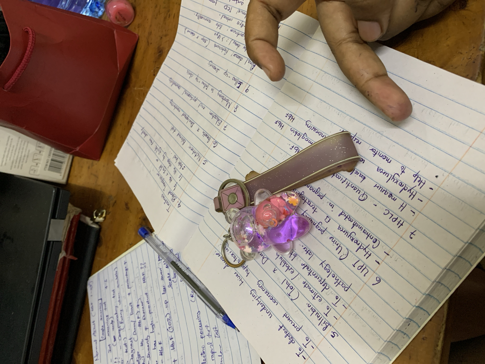 Here is a detailed description of the image:
**Scene Overview:**
The image presents a close-up view of a study or work area. There are handwritten notes on lined paper, a pen, a keychain, and a partially visible red gift bag. A hand is also visible in the frame.
**Specific Details:**
* **Handwritten Notes:** Two notebooks filled with handwritten notes are visible. The notes appear to be related to medical or scientific content, as terms like "Bilirubin," "HPLC," "Hydroxyurea," "Hemoglobin HbF," "Blood Transfusion," "Nutrition: Balanced diet," "Normal adult" "screening test" and others are discernible.
* **Pen:** A blue ballpoint pen rests on one of the notebooks.
* **Keychain:** A keychain with a pink and purple bear-shaped charm and a rectangular tag lies on top of the notes.
* **Hand:** A hand with dark spots on it is visible at the top right of the image, with the index finger pointing to the handwritten notes.
* **Red Gift Bag:** A red gift bag with a rope handle is positioned on the left side of the image.
* **Desk/Surface:** The objects are placed on a desk or surface with a dark finish.
**Contextual Clues (Tanzania, Dar es Salaam):**
Without additional information or context within the image itself, it is impossible to ascertain if this image was taken in Dar es Salaam, Tanzania. There are no recognizable landmarks, cultural cues, or other indicators to link the image to a specific location. The content of the notes does not provide any clues either.