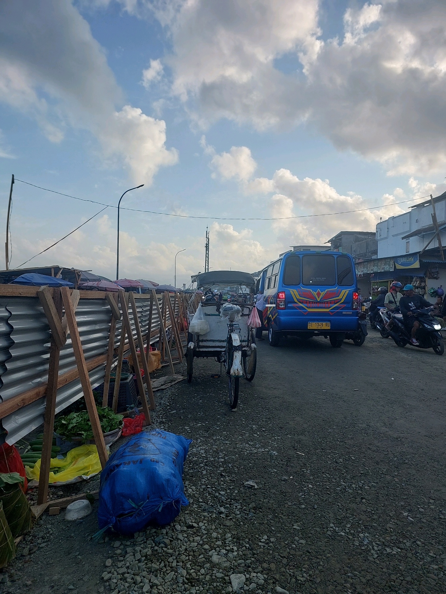 The image depicts a street scene in Ambon, Indonesia, likely at a market or roadside vending area. The foreground features a makeshift barrier constructed from corrugated metal and wooden supports, with various goods such as vegetables and produce displayed behind it. A large blue sack is prominently placed on the gravelly ground in front of the barrier.
Further into the scene, a three-wheeled bicycle, possibly a pedicab, is parked on the side of the road. Beyond that, a blue minibus with yellow and red decals is driving on the asphalt road, accompanied by traffic, including several motorcycles. One motorcycle is carrying two individuals, the rider wearing a helmet and the passenger also appearing to have headwear. Another motorcycle is visible further back in the traffic.
In the background, a row of buildings and stalls suggests a commercial area. A tall tower, possibly a telecommunications tower, is visible in the distance. The sky is partly cloudy with patches of blue. The overall impression is of a busy, outdoor market environment during daylight hours.