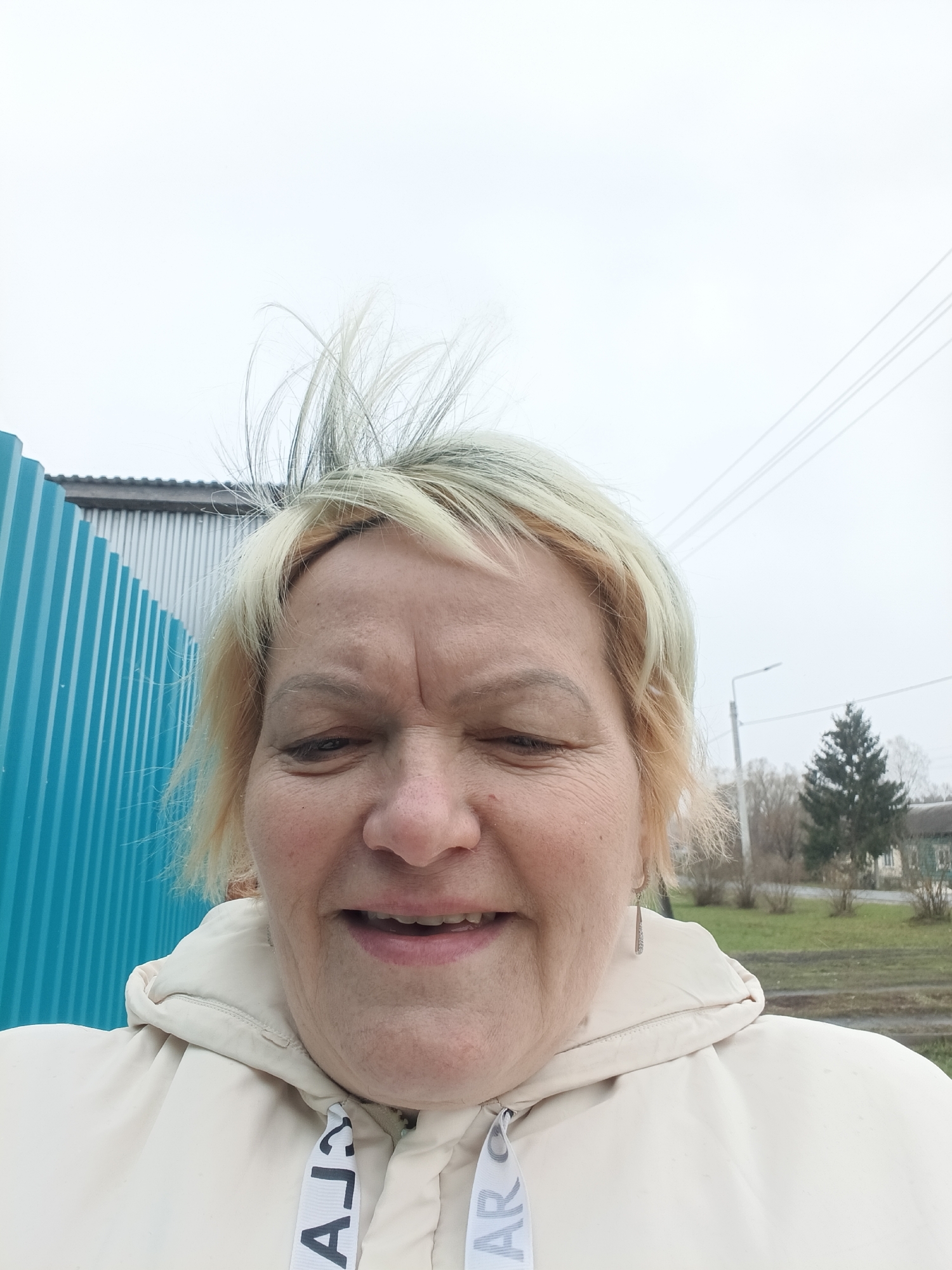 A close-up selfie features a woman with blonde hair against an overcast sky. Her hair is somewhat disheveled, with some strands standing upright. She is wearing a light-colored hooded jacket with a drawstring.
In the background, a blue corrugated metal fence is visible on the left. Further back, a building with a corrugated metal roof is partially visible. A street light and a small pine tree are visible in the distance, along with a low-lying building. The scene appears to be in a rural or suburban area. The location is Красный Маяк, Russia.