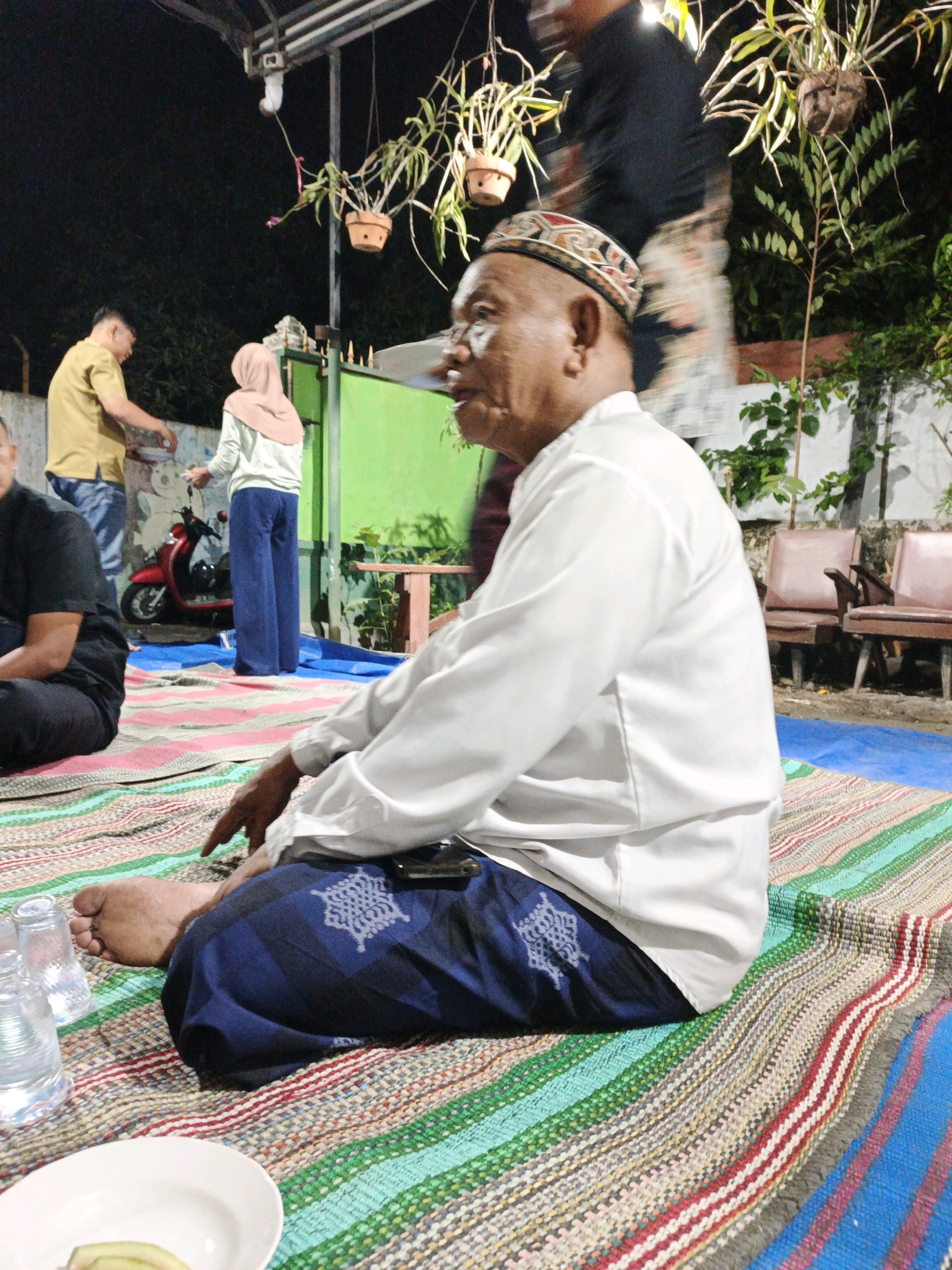In Sumber Gedang, Indonesia, at night, an older man is seated on a colorful woven mat. He is wearing a white long-sleeved shirt, a dark blue sarong with a white patterned design, and a patterned cap. His left foot is visible, resting on the mat. To his left, there are two clear plastic cups and a white plate with a slice of green fruit. The background shows other people in casual attire and a red scooter. Plants hang in pots from an overhead structure, and in the distance, two chairs are visible. The scene appears to be an outdoor gathering.
