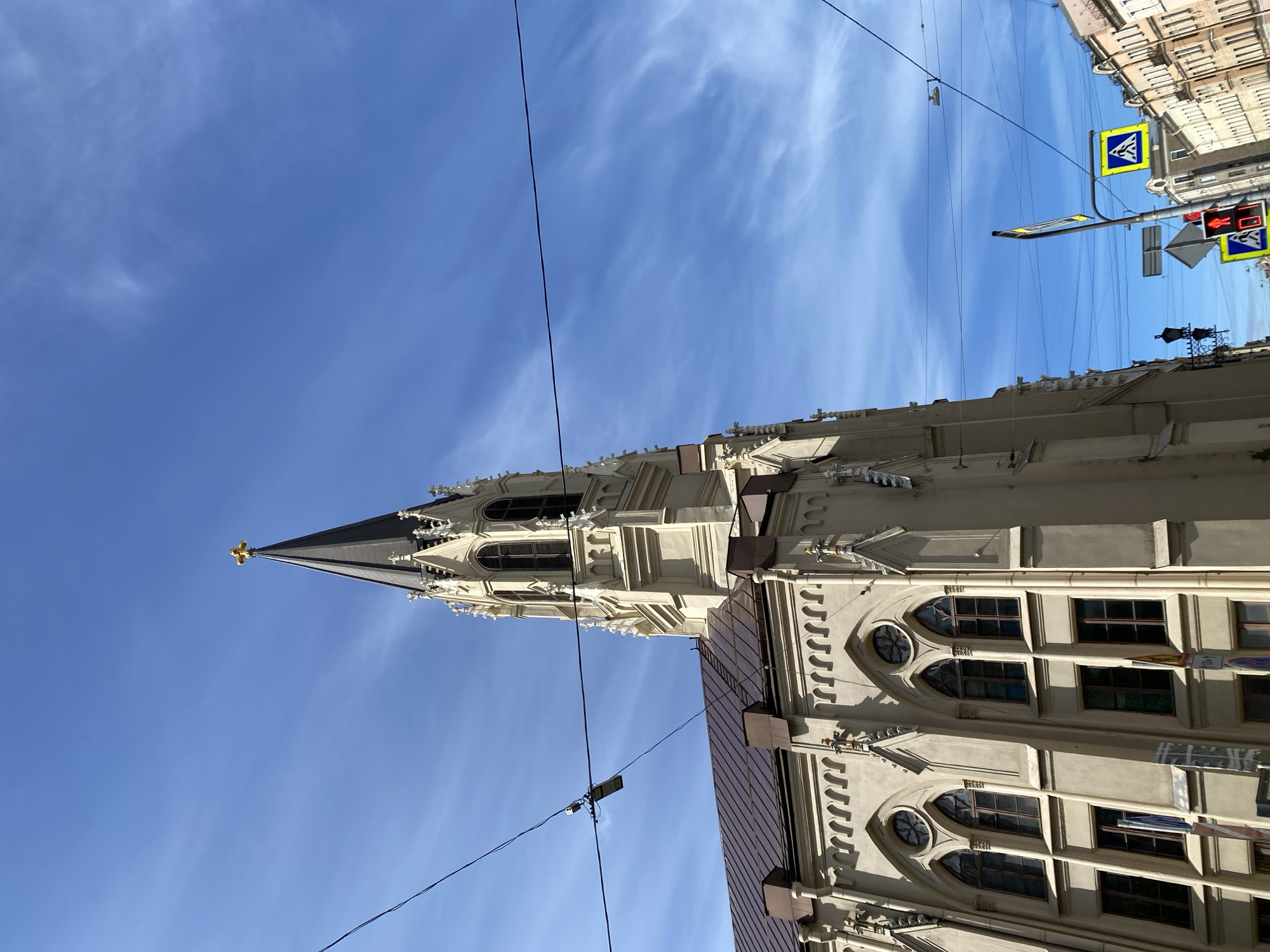 The image depicts a tall, light-colored Gothic-style building, possibly a church or cathedral, in Saint Petersburg, Russia. The building has a sharp spire with a golden cross at the top and is decorated with intricate stone carvings, pointed arches, and numerous windows. In the foreground, there are dark power lines running diagonally across the image. To the right of the building is a pedestrian crossing sign, and a red traffic light is also visible. The sky is clear blue with wispy white clouds.