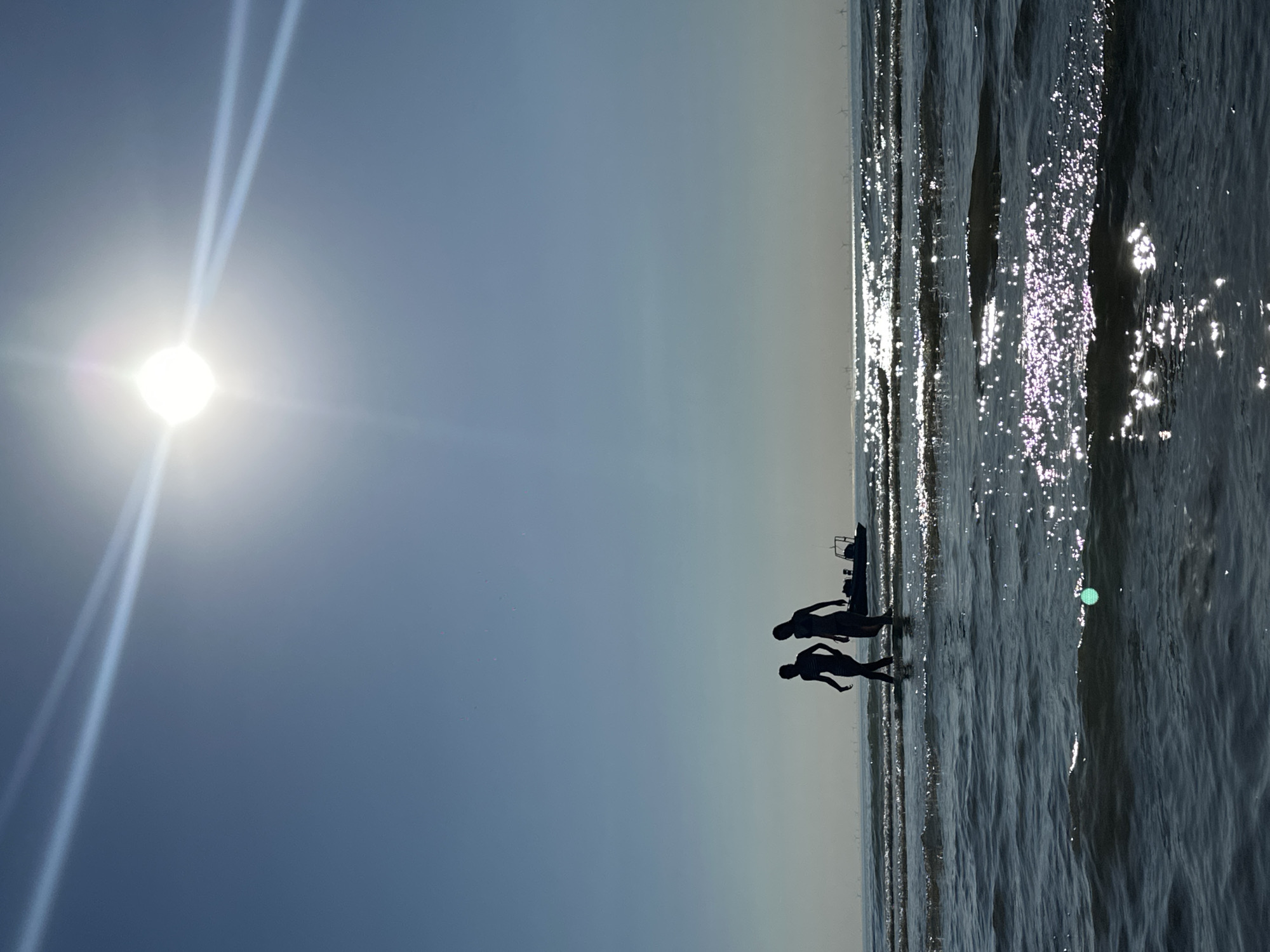 Two individuals, silhouetted against a bright sun and clear blue sky, walk in shallow water at the beach in Zandvoort, Netherlands. A small boat is visible to the right of the individuals. The sun's glare creates bright reflections on the surface of the water, which has small waves. In the distance, a line of wind turbines is visible on the horizon. The overall scene is one of a bright, sunny day at the seaside.