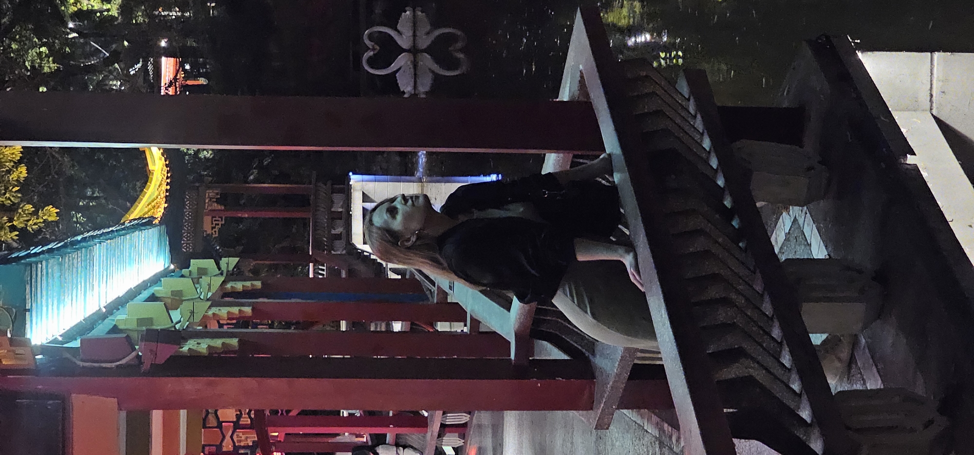 A woman with long hair is shown at night. She is standing on a red and brown wooden structure. In the background there is an illuminated structure which appears to be a Chinese-style building with a blue tiled roof, some of which is illuminated with yellow lights. A heart-shaped sculpture is visible in the upper part of the image. The location appears to be in the city of Yuzhong District, China.