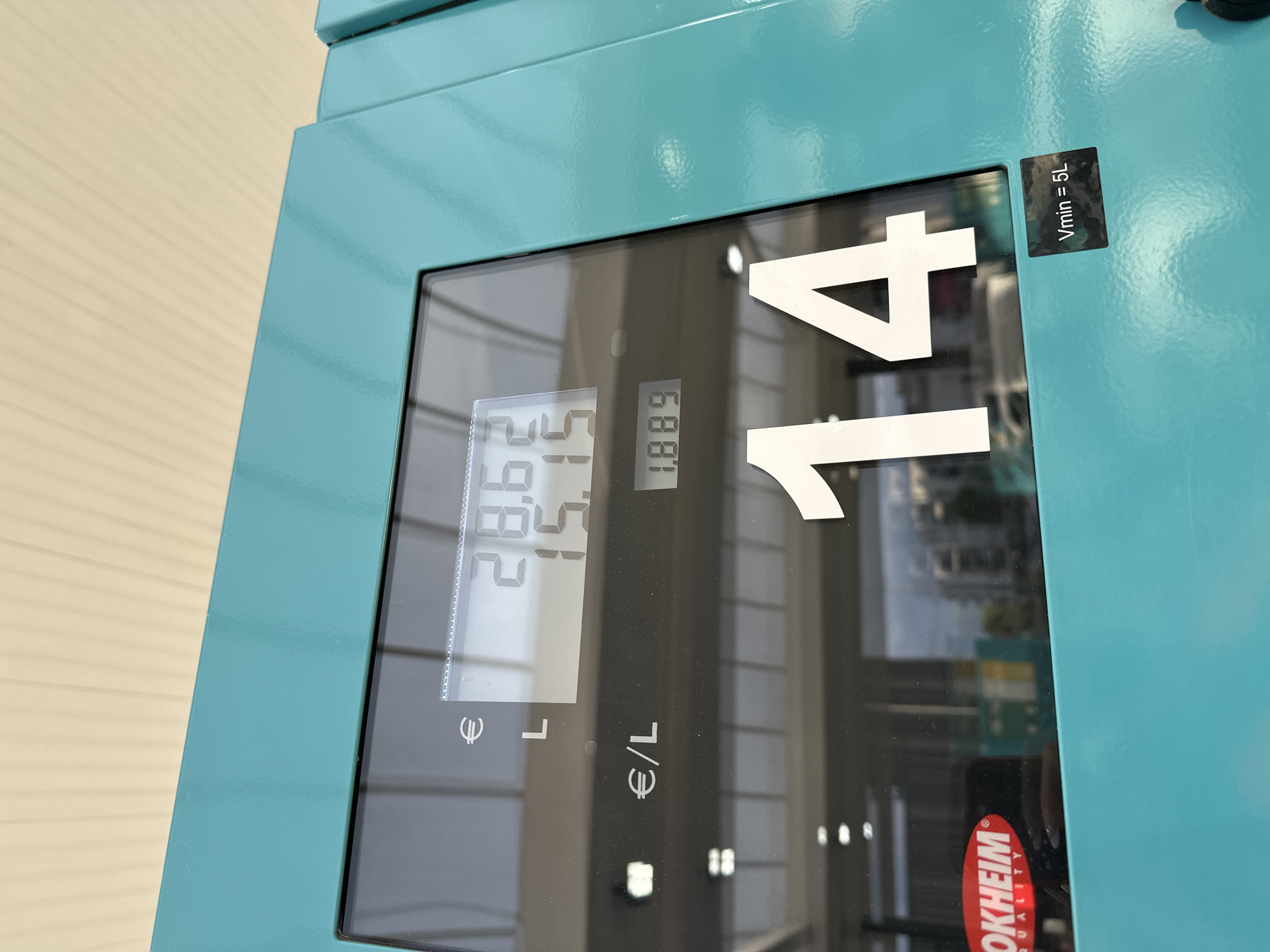 The image shows a close-up of a digital display on a turquoise fuel pump, likely at a gas station. The display shows a large number "14" in white text, signifying the fuel quantity in liters. Above this, another display shows "€/L" next to a price of "1.889". Higher still, another display shows "€" and "L" with corresponding values of "28.62" and "15.". A red logo with the text "OKHEIM QUALITY" is visible on the left side of the pump. A small black sticker at the bottom right of the pump reads "Vmin = 5L". The background suggests an outdoor environment with overhead structures and reflections of buildings and sky in the pump's glass. The location is identified as Wasserbillig, Luxembourg.