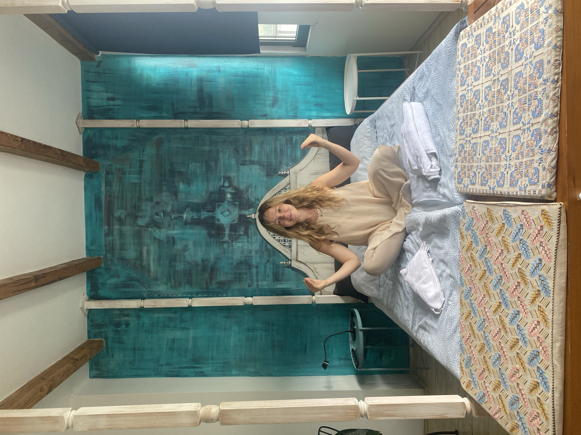 A woman sits cross-legged on a bed in a bedroom. The bed has a white headboard and posts, and is covered with a blue and white patterned duvet. Two white towels are folded neatly on the duvet. A woman with blonde hair, wearing a light-colored dress, is sitting on the bed with her legs crossed and her hands raised. She is looking at the camera and smiling. Behind the bed is a wall painted with a teal and black abstract mural. There are wooden beams on the ceiling above the mural. To the left of the bed is a small, round, teal-colored nightstand with a black lamp. To the right of the bed is a small, round, white side table. The floor appears to be made of wood planks. The overall style of the room is bohemian and eclectic, suggesting a hotel room or a guest house in Belgrade, Serbia.
