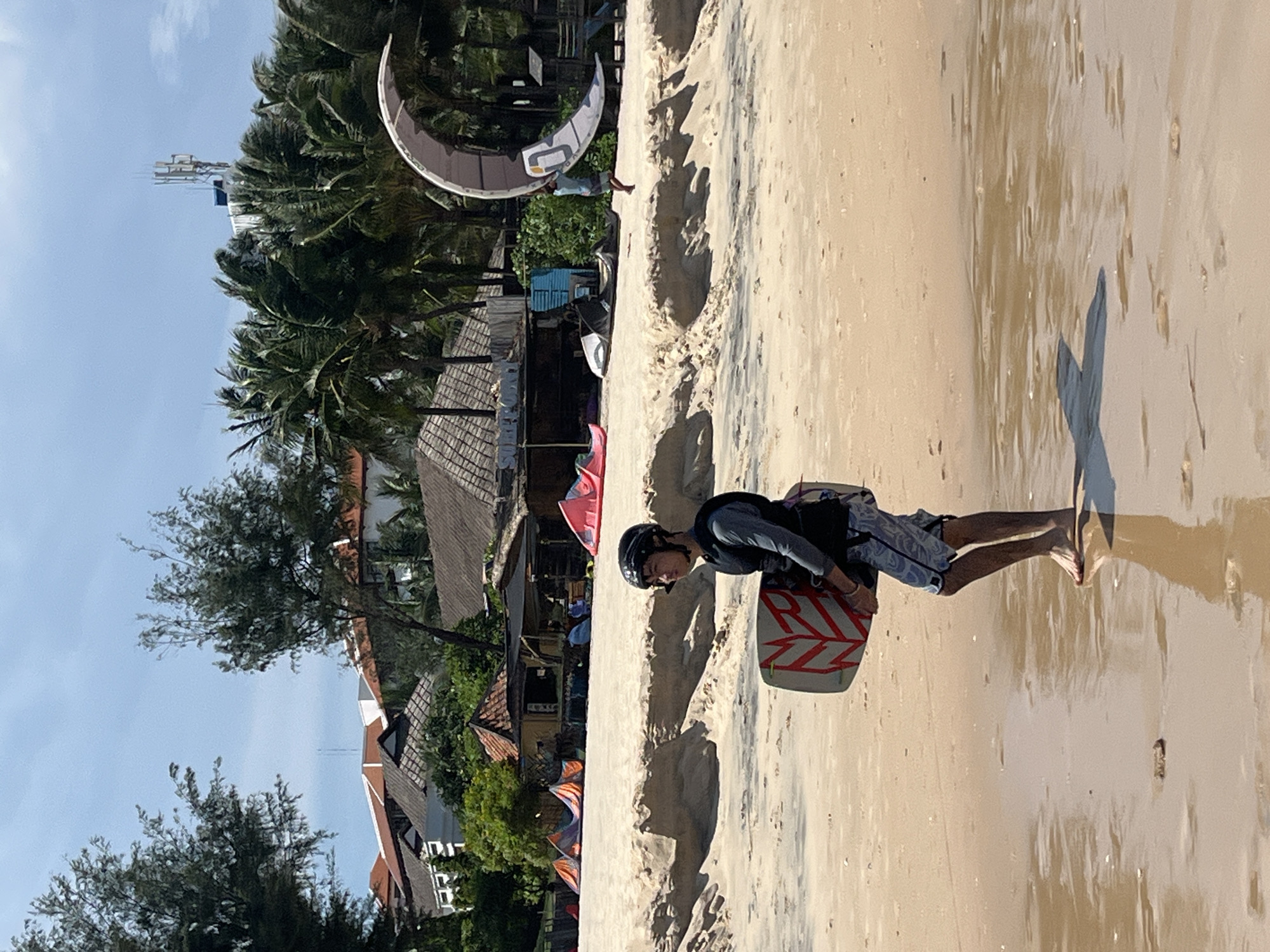 A young man in a helmet and life vest walks on a wet sandy beach in Phu Thuy Ward, Vietnam, carrying a kiteboard. The kiteboard is white with red geometric patterns. He is wearing patterned shorts. The beach has footprints and small puddles reflecting the sky. In the background, there are several kites set up on the sand, small buildings with thatched roofs, palm trees, and a large crescent-shaped kite in the air. A cell tower is visible on a building in the distance. The sky is clear and blue with some clouds.