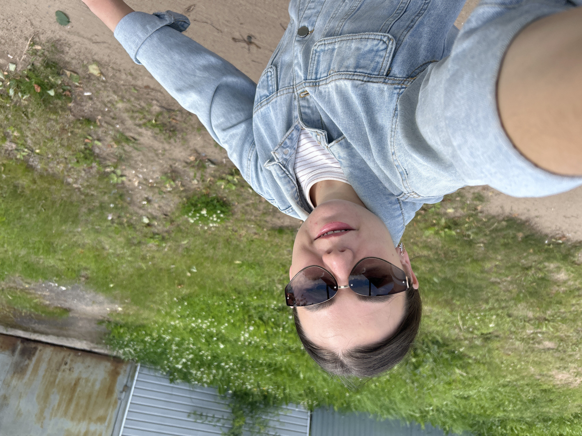 A woman is taking a selfie outdoors. She is wearing a light blue denim jacket over a striped t-shirt and large, dark-lensed sunglasses. The woman has dark hair pulled back and is smiling slightly. She is standing on a dirt path, with green grass and small white flowers on either side. In the background, a light-colored metal building with a large, rusted metal door is visible. The scene appears to be in a rural or industrial area, consistent with the outskirts of Chaikovsky, Russia, possibly near a dacha or garage. The lighting suggests it is daytime, with diffused sunlight.