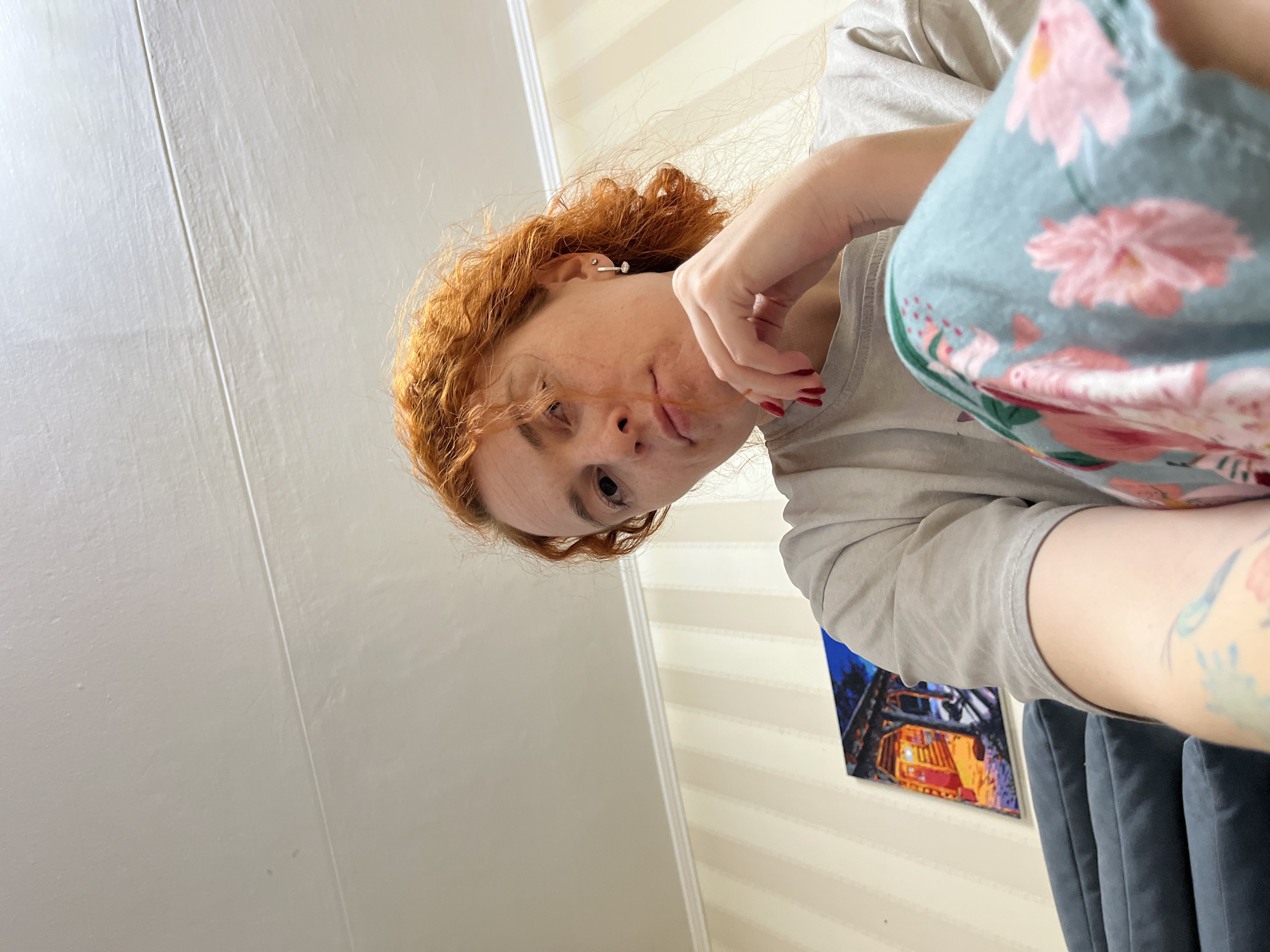 A woman with red curly hair, wearing a light grey t-shirt and light blue floral pants, looks down at the camera. She has red nail polish and a small stud earring. In the background, a white door is to the left and horizontally striped wallpaper to the right. A painting of a structure with bright red and yellow lights hangs on the wall and a dark blue velvet chair is visible to the right of the painting. The location is implied to be in the city of Chaikovsky, Russia.