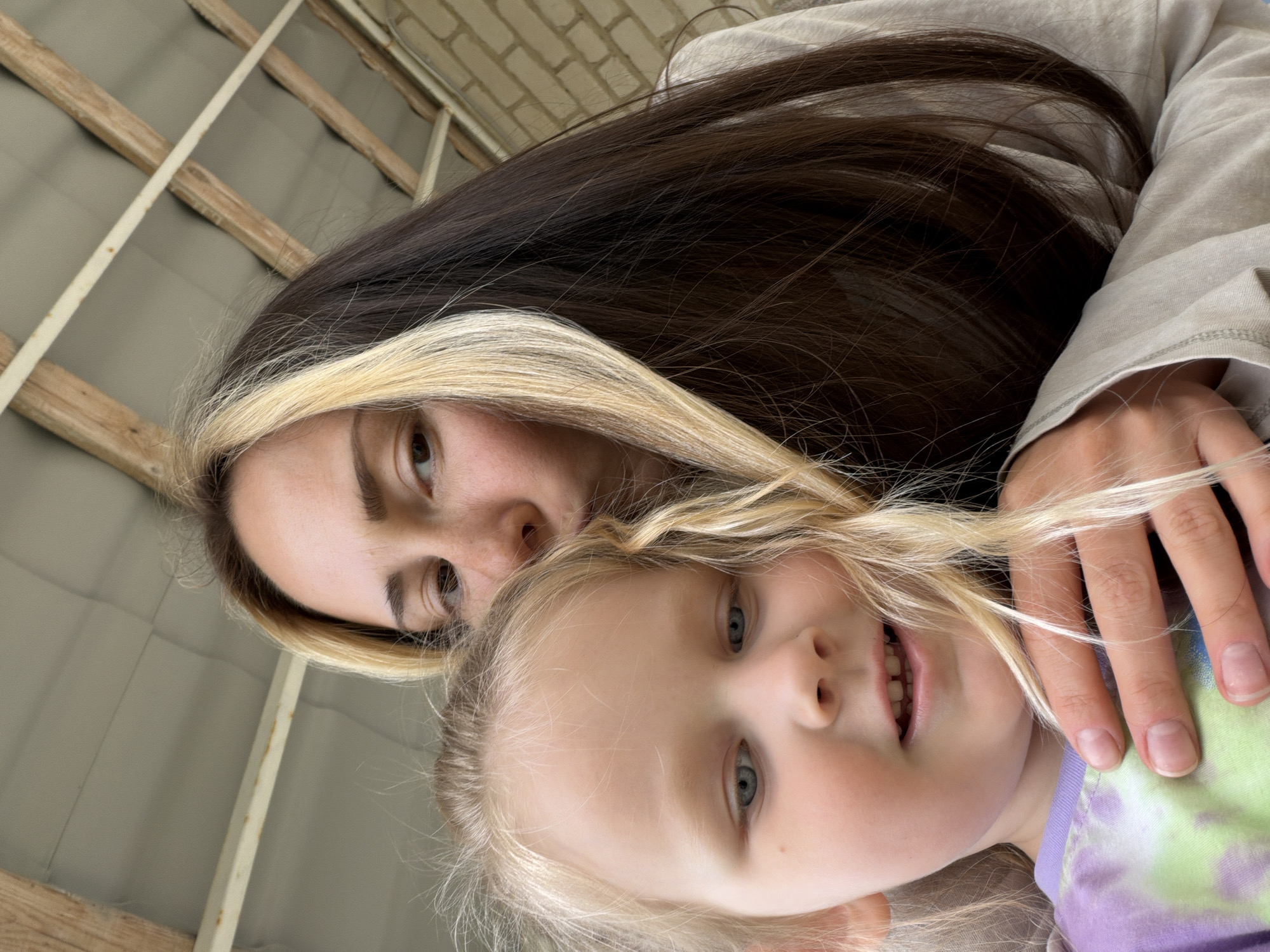 A woman and a young girl are looking down at the camera. The woman has brown hair with a blonde streak and is wearing a light grey shirt. The girl has blonde hair and is wearing a purple and green tie-dye shirt. A hand is visible near the girl's face. The background shows a ceiling with wooden beams and grey panels. There is no indication this photo was taken in Kamyshin, Russia.