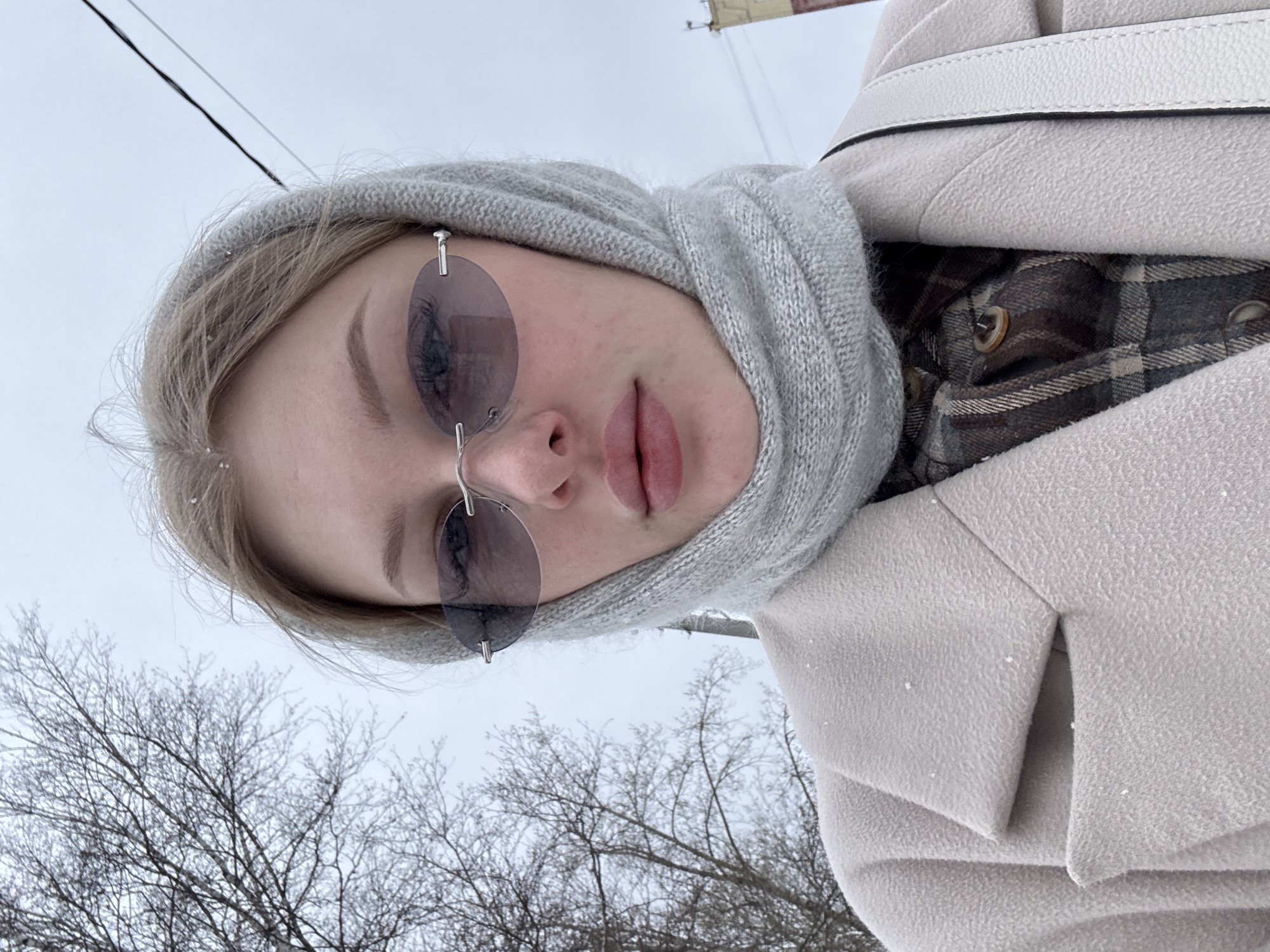 A vertical, close-up selfie captures a young woman outdoors in a winter setting. The woman, with light hair, looks directly into the camera with a neutral expression. She wears stylish, rimless sunglasses with dark, oval-shaped tinted lenses, which obscure her eyes. Her lips are full and have a natural pink hue.
She is dressed for cold weather, wearing a light grey knitted headscarf or hood that covers her head and frames her face. She has on a light beige or off-white textured winter coat, and a white strap, likely from a shoulder bag, is visible over her right shoulder. A glimpse inside her coat reveals a brown and grey plaid shirt with buttons. Small white flakes, consistent with light snow or frost, are scattered on her hair and coat.
The background features a stark, overcast sky and the intricate, leafless branches of deciduous trees, typical of a winter landscape. These branches appear to be lightly dusted with snow or frost. A thin black utility wire or cable is visible across the top right portion of the frame.
The image portrays a serene, personal moment on what appears to be a cold, subdued winter day in Perm, Russia. The soft, diffused lighting and muted color palette contribute to a calm atmosphere. No discernible text is visible in the image.