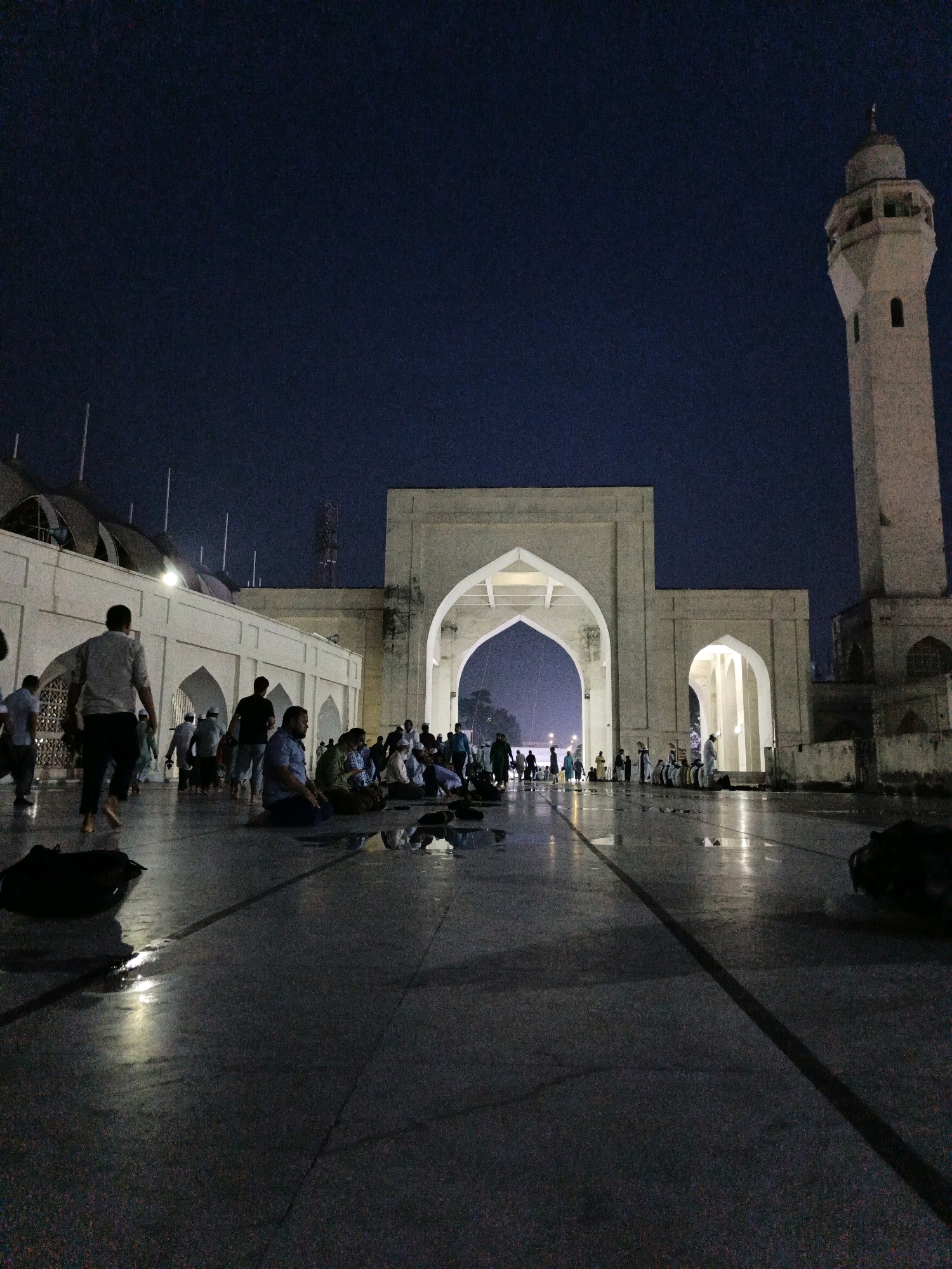 This image depicts a nighttime scene at the Baitul Mukarram National Mosque in Dhaka, Bangladesh. A group of people are gathered in the open courtyard, many of whom are seated on the ground, some in prayer. The courtyard floor is wet, reflecting the lights from the mosque and creating puddles. The mosque itself features a tall, white minaret on the right side of the frame and a large, arched entrance in the center. People are seen walking and standing near the entrance, while others are positioned further into the courtyard. The sky is dark, indicating it is nighttime, with minimal ambient light visible. The overall atmosphere suggests a religious gathering or prayer service.