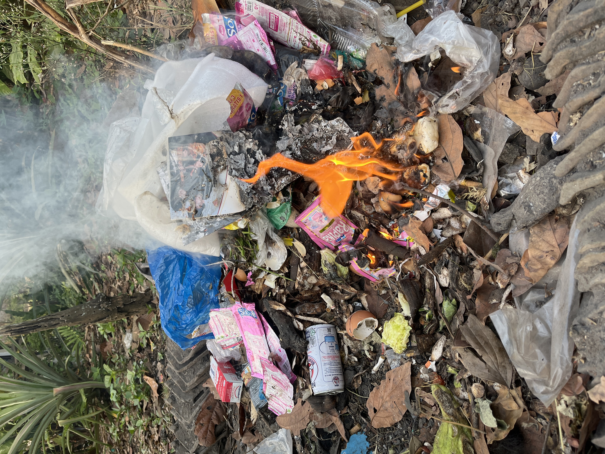 The image depicts a pile of burning trash outdoors, likely in a natural or semi-natural environment, given the presence of dry leaves, vegetation, and what appears to be discarded building materials. The fire is visible in the center of the frame, with flames reaching upwards and emitting smoke that drifts into the air. Among the burning debris are various forms of waste, including plastic bags, cardboard packaging, and what seems to be a crushed aluminum can. Some of the packaging is brightly colored, suggesting consumer goods. In the background, there are large green plants, possibly a pineapple plant, and trees, indicating an outdoor setting that is not a formal city street but could be on the outskirts of Kebumen, Indonesia, in a more rural or less developed area. The ground is covered with fallen leaves and dirt, reinforcing the outdoor, potentially unkempt environment. There are no people visible in the image.