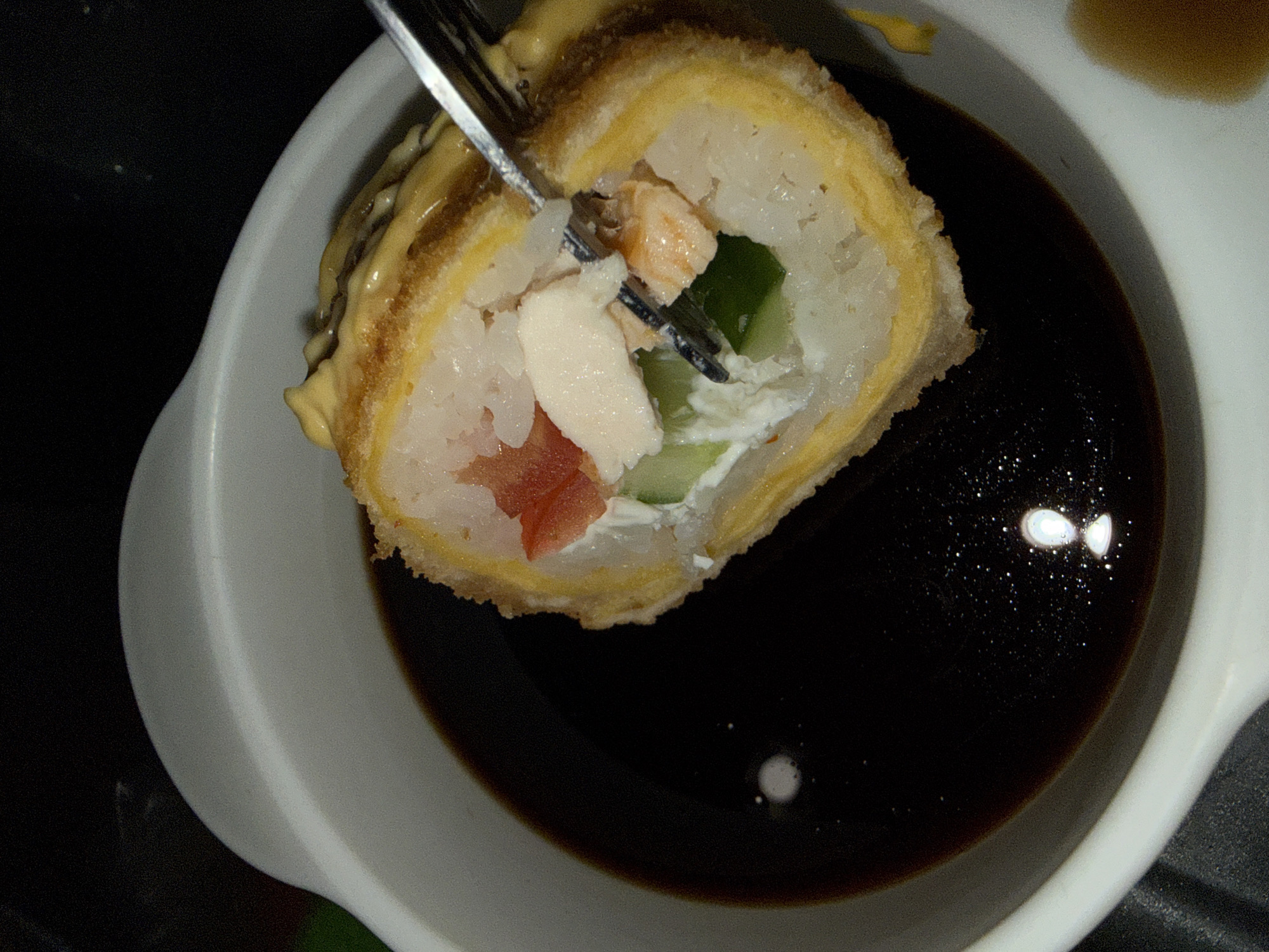 A partially consumed sushi roll, impaled by a silver fork, sits above a bowl filled with a dark liquid, possibly soy sauce. The sushi roll appears to be topped with a light-colored sauce. Inside, rice surrounds visible ingredients including tomato, cucumber, and seafood such as salmon and white tuna or crab. The roll has a fried batter coating. The scene is set in Dmitrov, Russia.
